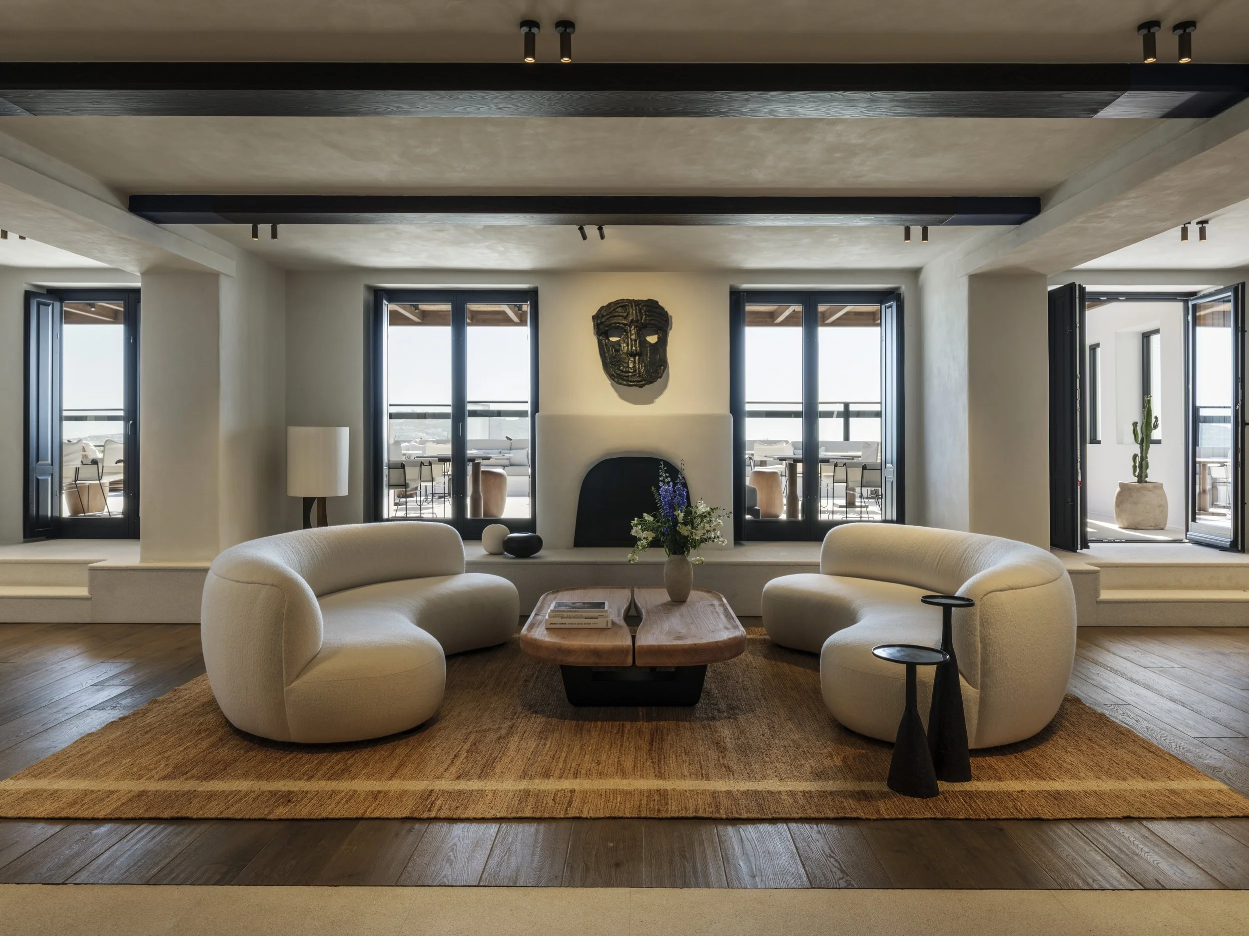 Living room with beige curved sofas, a wooden coffee table with books and flowers, a large area rug, black side tables, modern decor, and large windows with black frames overlooking a balcony with outdoor furniture and a cactus.