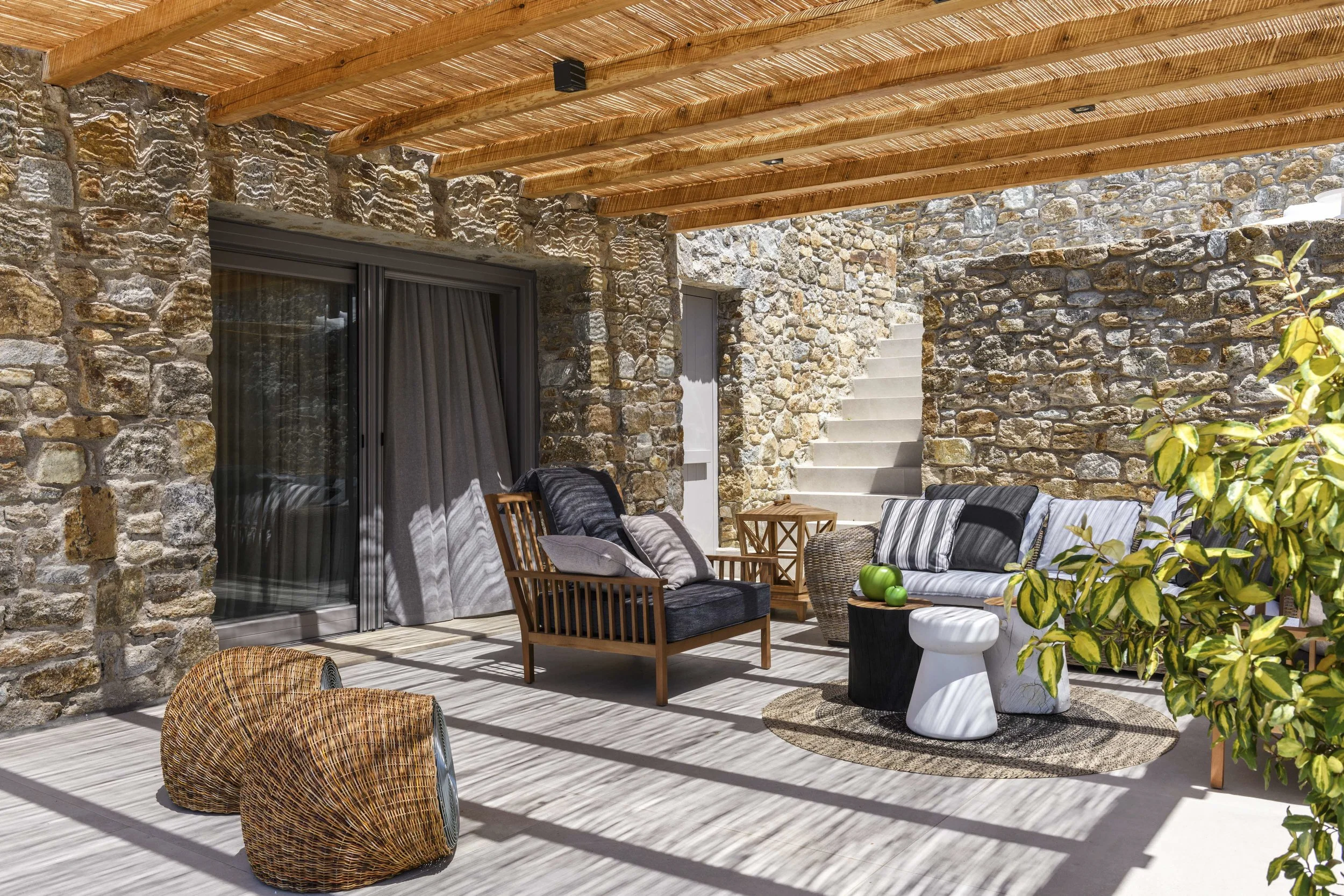 Outdoor patio with stone walls and wooden ceiling, furnished with wicker and wooden chairs, sofas with striped cushions, a small round wooden table, a white planter with a green plant, and a rug, partially shaded by a tree.