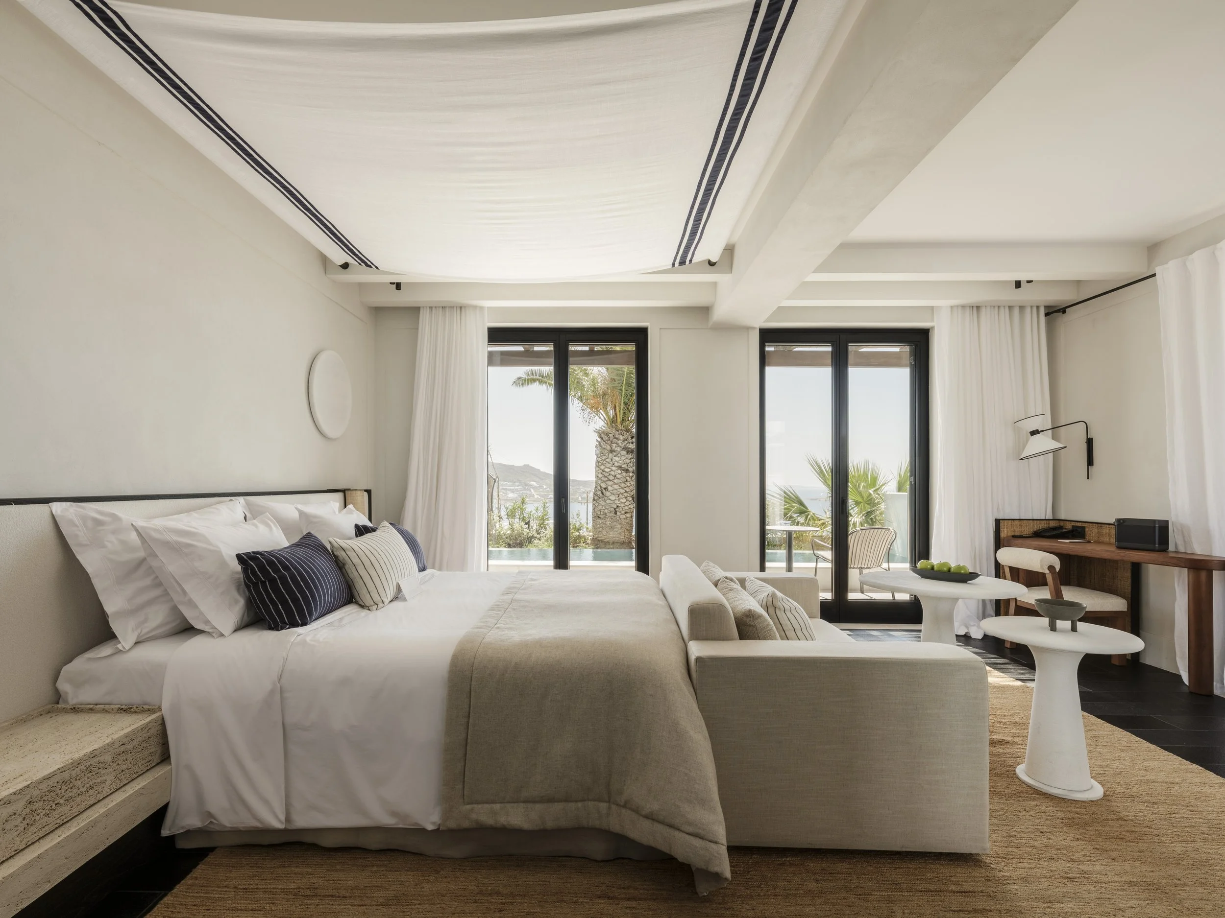 A modern bedroom with a bed covered in white linens and striped pillows, a beige sofa at the foot of the bed, large glass doors with white curtains, and a view of palm trees and a mountain outside.
