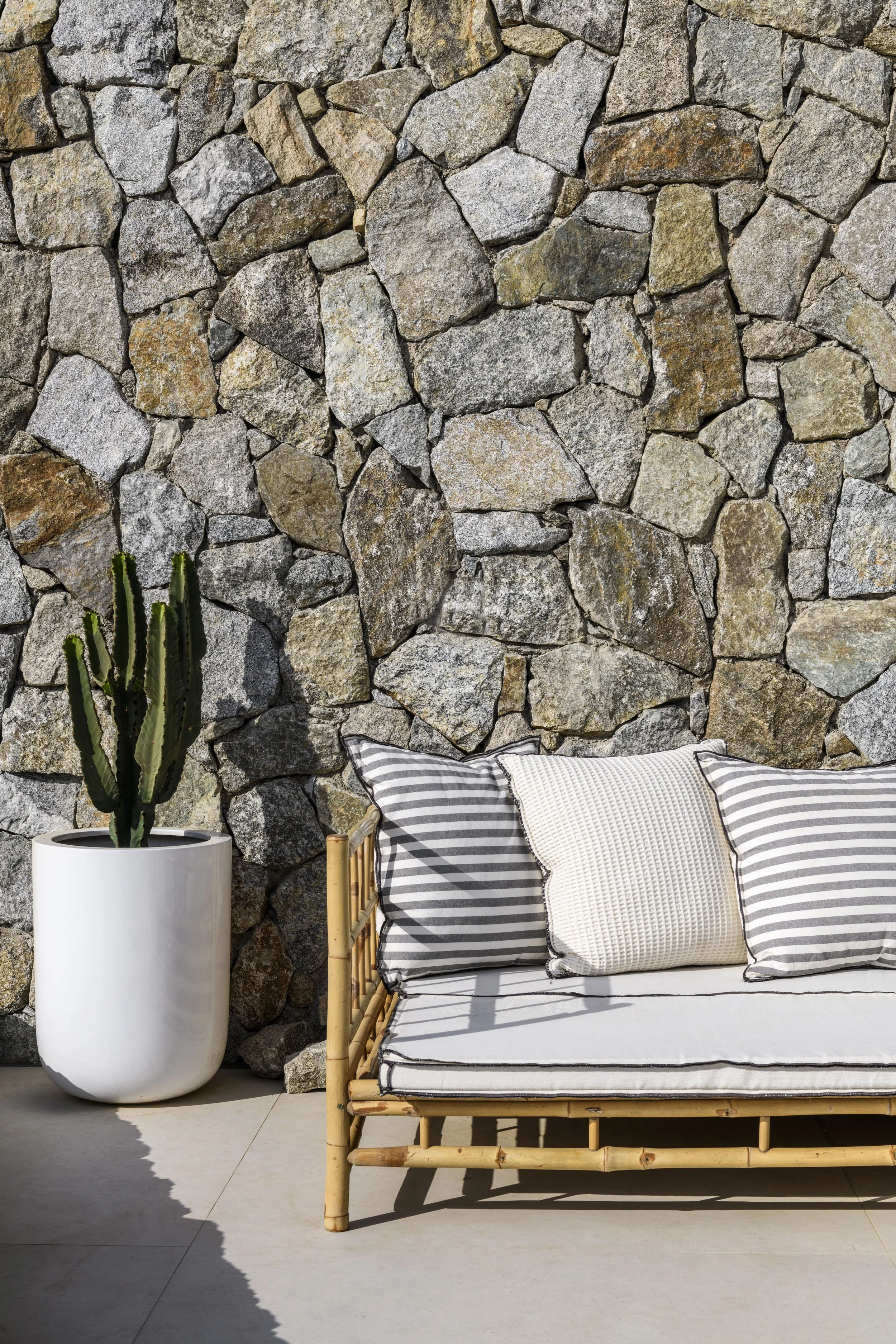 A stone wall with large rocks, a green cactus in a white pot, and a bamboo sofa with striped pillows and a beige cushion.