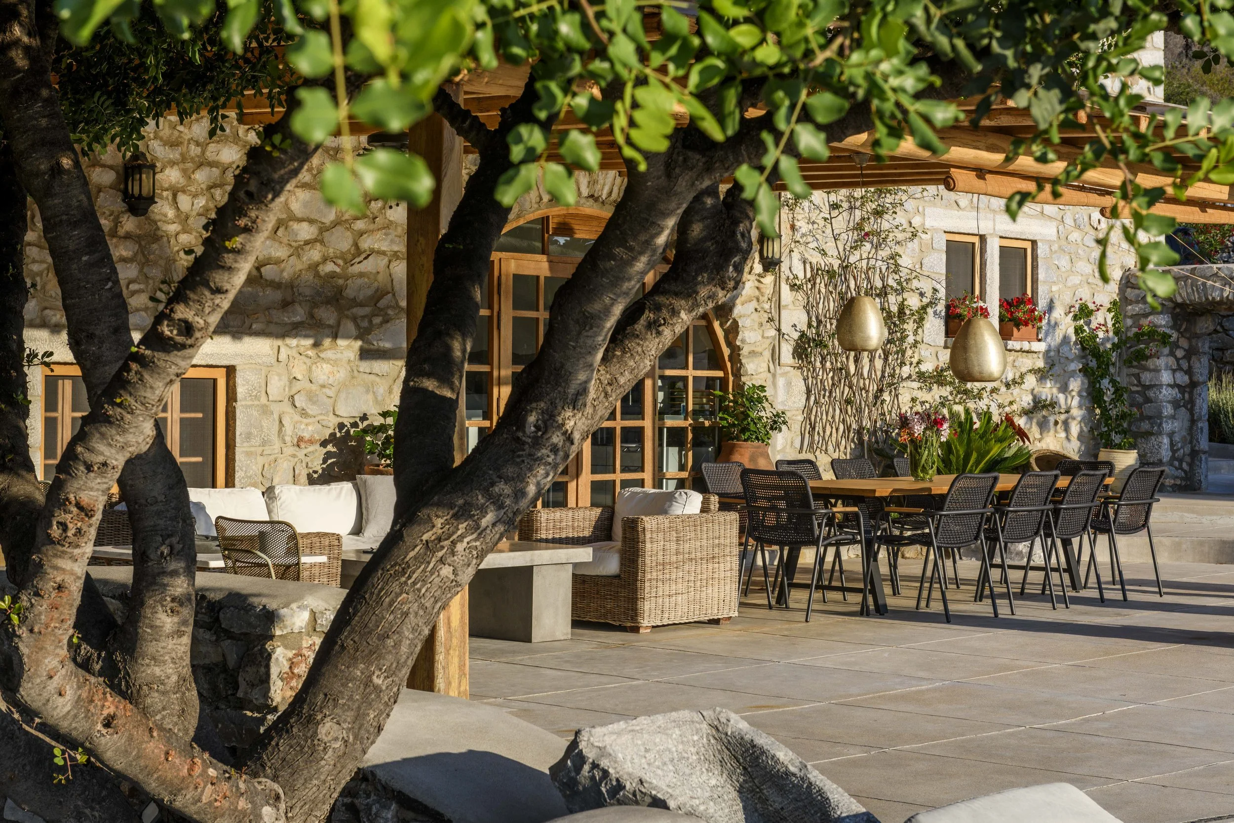 Outdoor patio area with stone building, wooden beams, trees, potted plants, and black outdoor furniture.