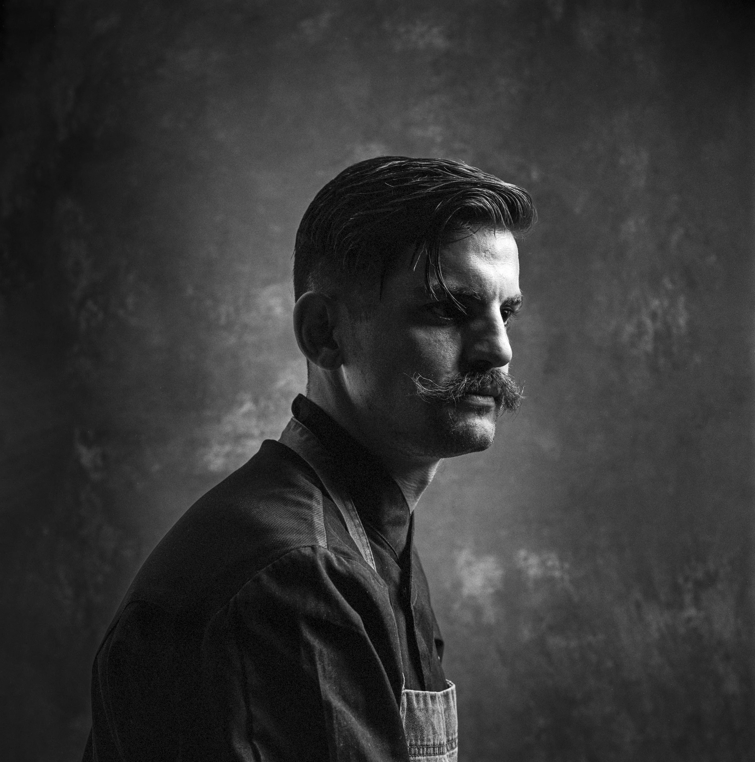 Black and white portrait of a man with neatly styled hair, a beard, and mustache, looking to the side, wearing a jacket, against a textured dark background.