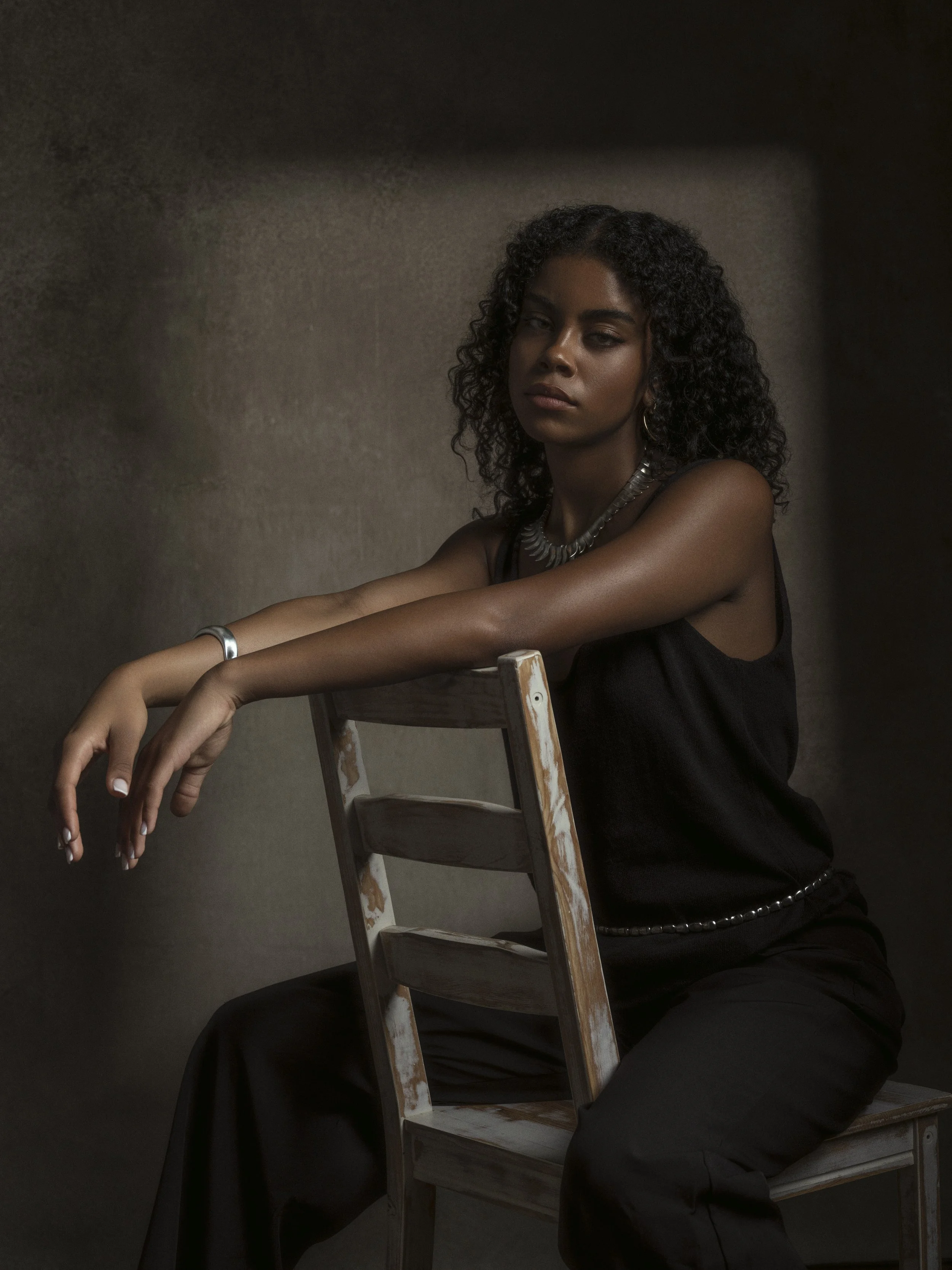 A young woman with curly hair wearing a black top and silver jewelry, sitting on a chair in front of a dark, textured background.