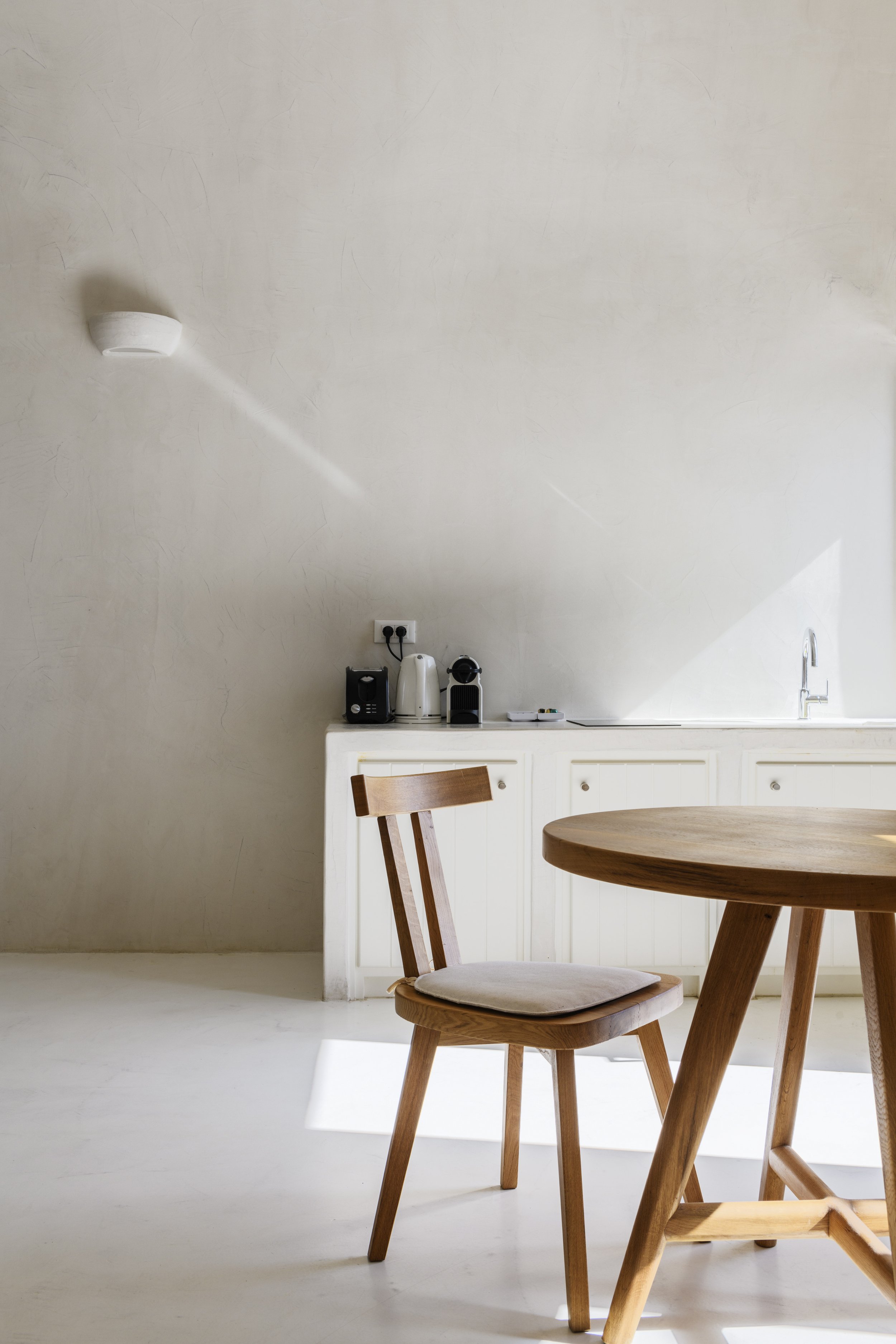 A minimalist kitchen with white walls, a wooden dining table, and matching wooden chairs. There are coffee machines on the counter and sunlight streaming in.