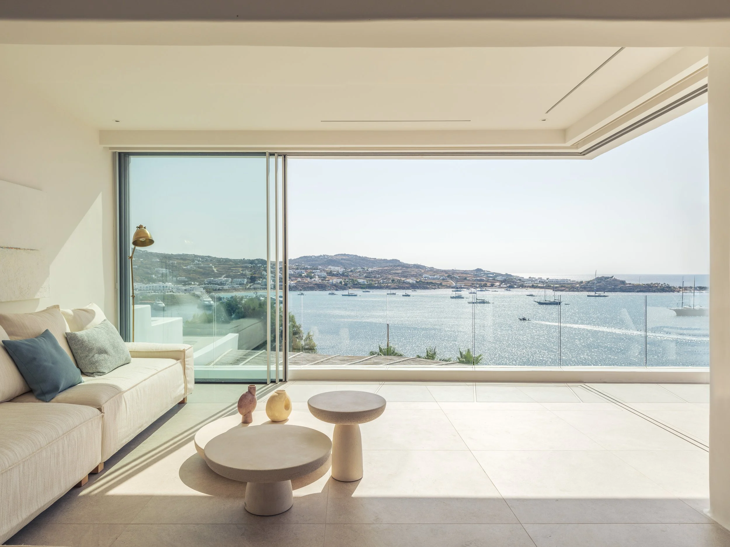 Modern living room with large floor-to-ceiling windows overlooking a harbor with boats and distant hills, minimalistic furniture, and decorative vases on a round coffee table.