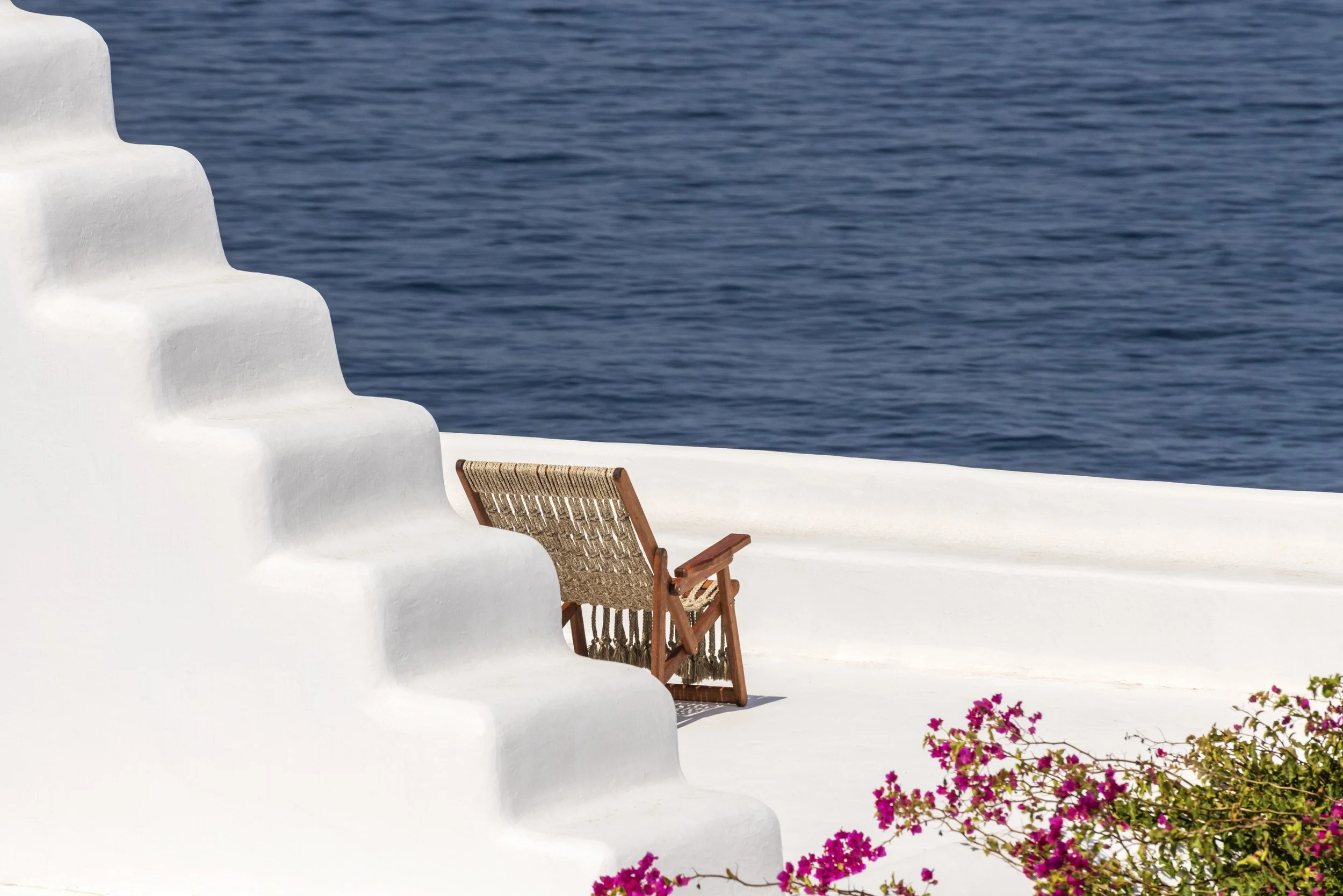 Minimalist white terrace of a luxury Mykonos island villa, featuring a traditional woven lounge chair and vibrant pink bougainvillea overlooking the deep blue sea.