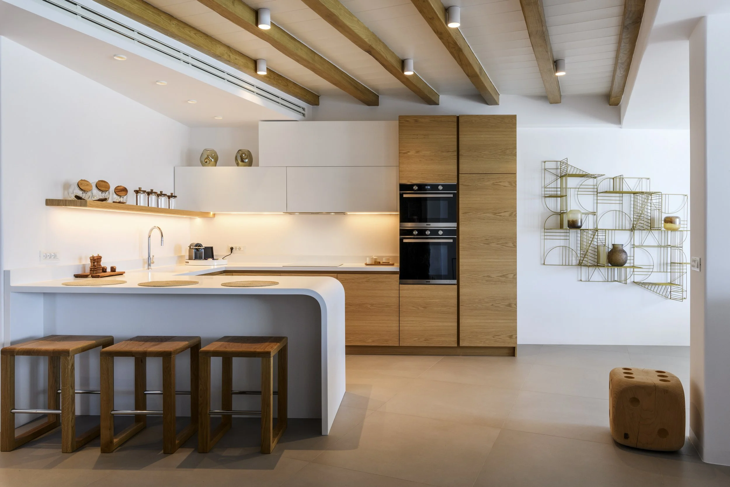 Modern minimalist kitchen with white countertops, light wood cabinets, gold wire wall shelves, and four wooden bar stools at a white kitchen island.