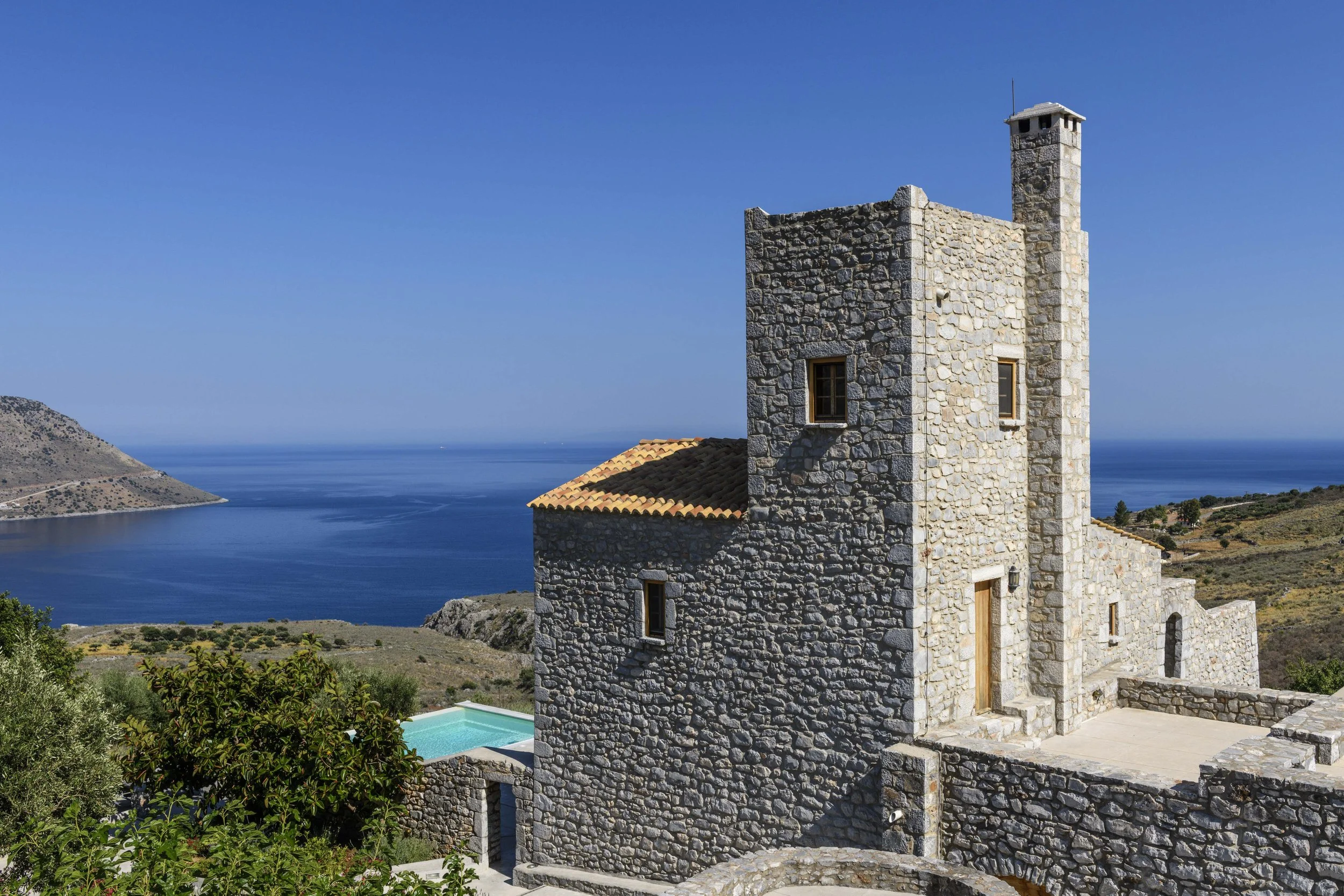 A stone building with a small tower overlooking a blue body of water and coastal hills under a clear blue sky.