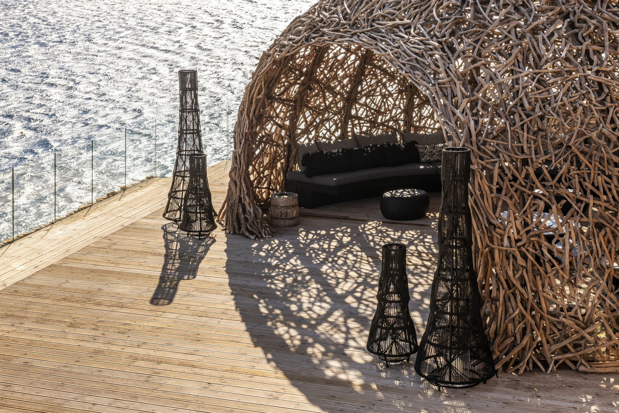 A seaside wooden deck with a large woven driftwood cabana and black seating area, surrounded by decorative black lanterns, with water reflecting sunlight in the background.