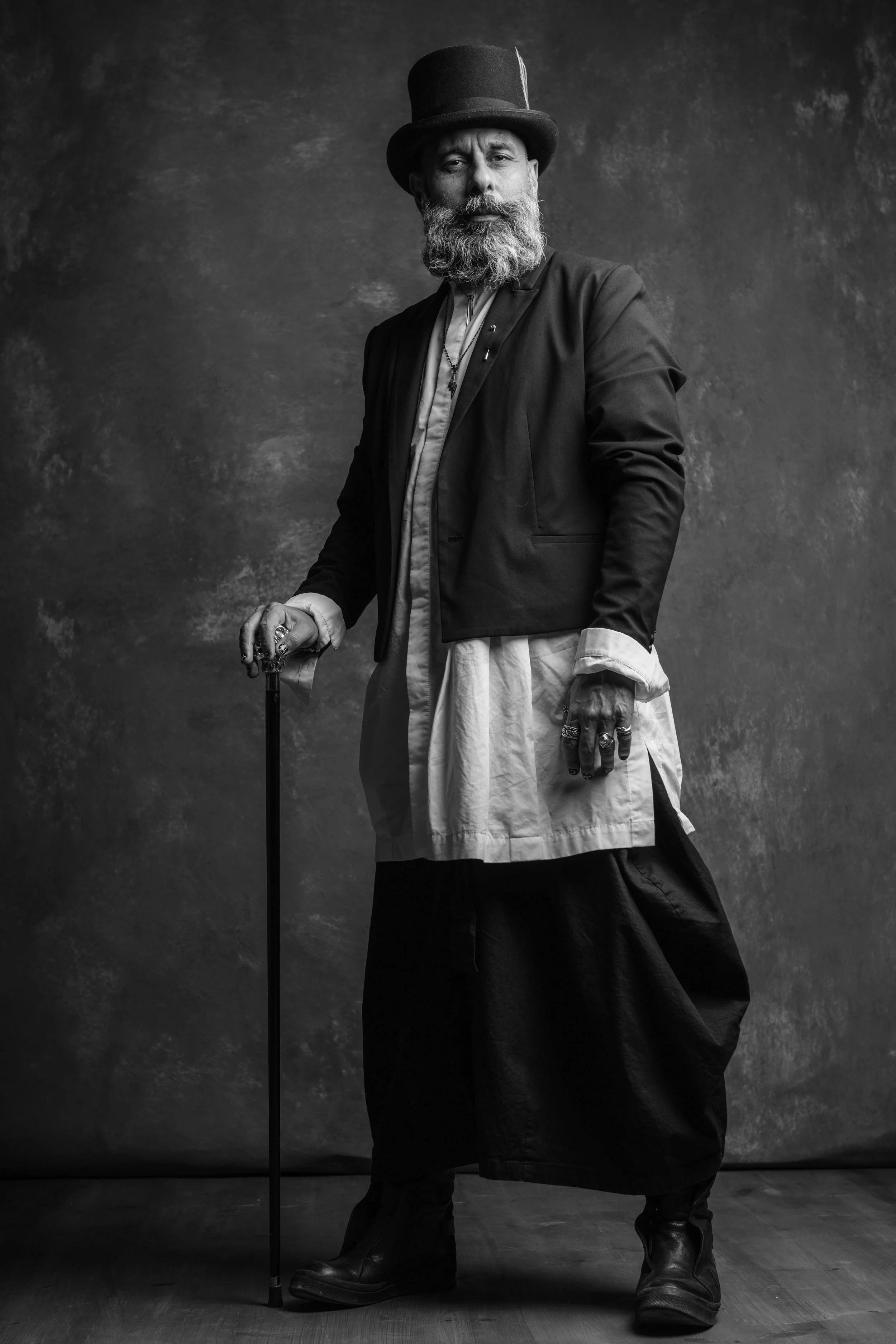 A black and white portrait of an older man with a long beard, wearing a top hat and a layered outfit, standing with one hand on a cane and the other on his side, in front of a plain textured background.