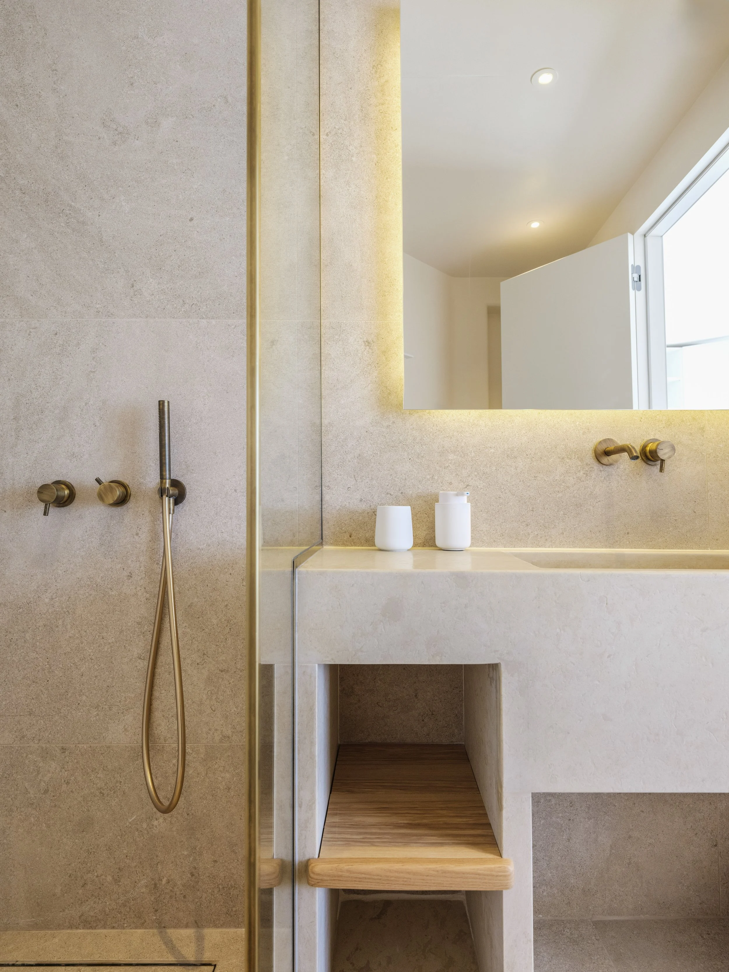 Modern bathroom with beige tiled walls, a large mirror, and a wooden shelf under the sink. The bathroom features minimalistic design with golden fixtures and a skylight window.