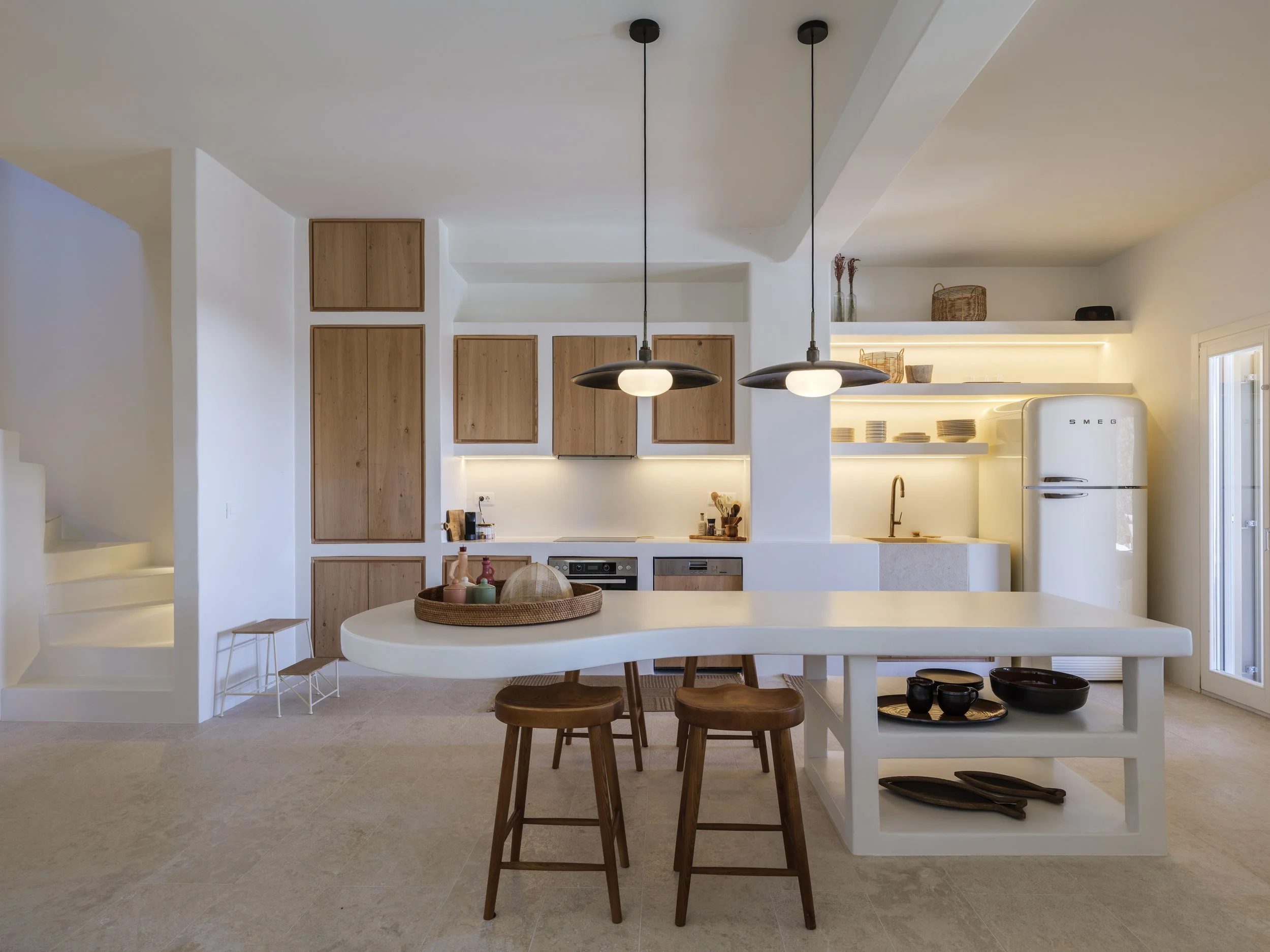 Modern kitchen with white walls, wooden cabinets, a white island table with wooden chairs, and black pendant lights hanging above.