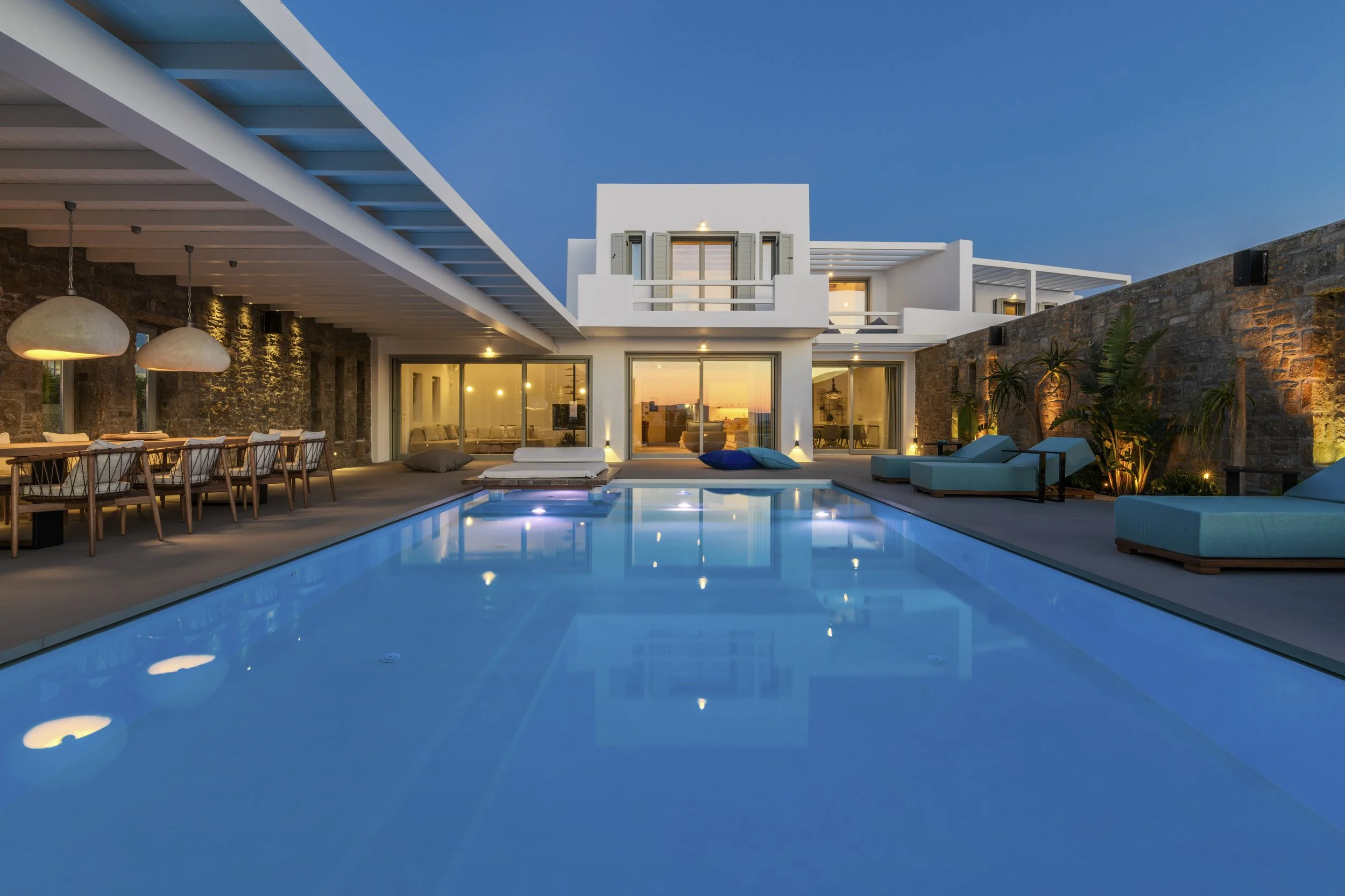 Modern house with large glass doors and outdoor pool area during sunset, illuminated with warm lighting, featuring seating and tropical plants.