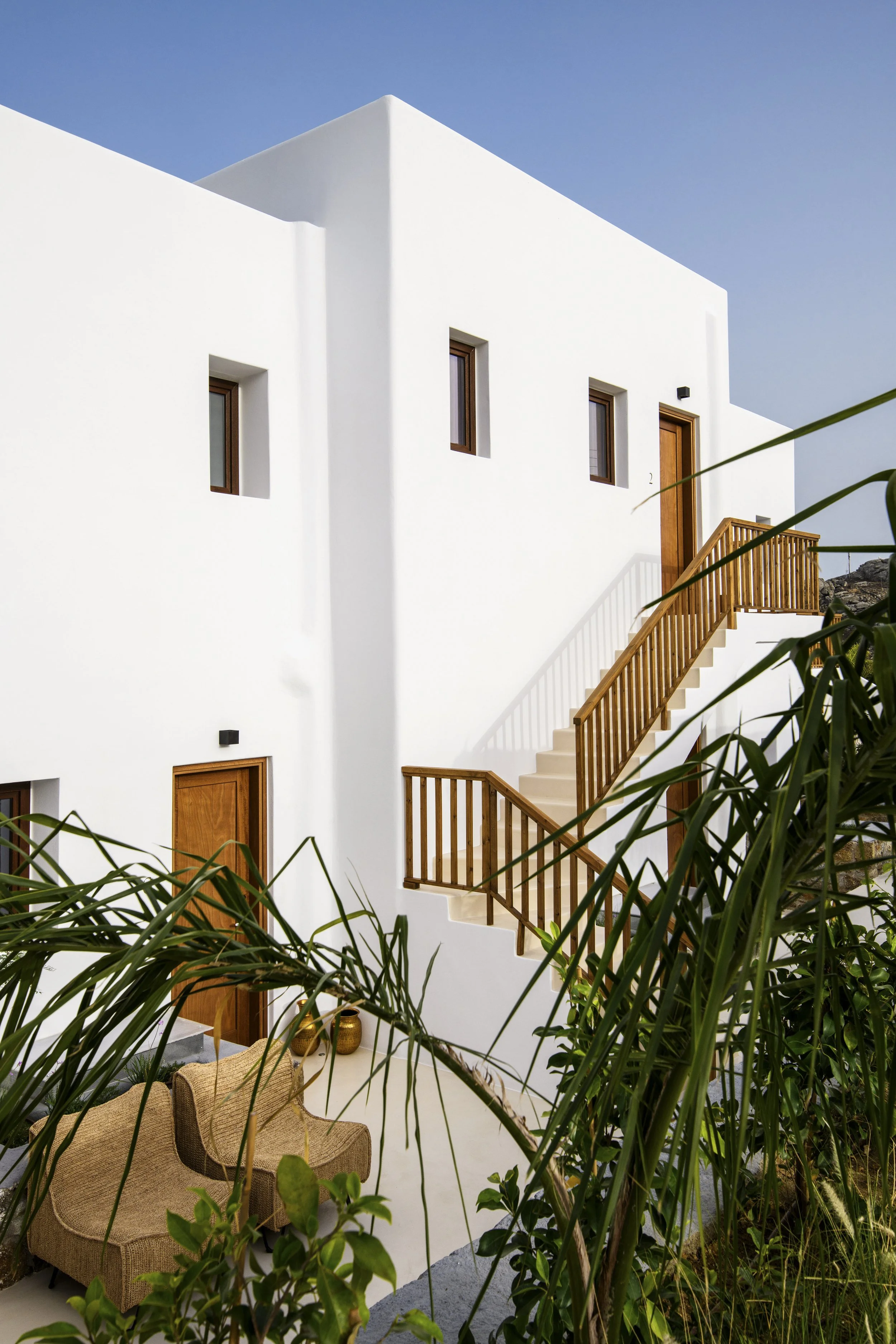 White modern building with small rectangular windows, wooden stairs, and a porch, partially obscured by green foliage in the foreground, under a clear blue sky.
