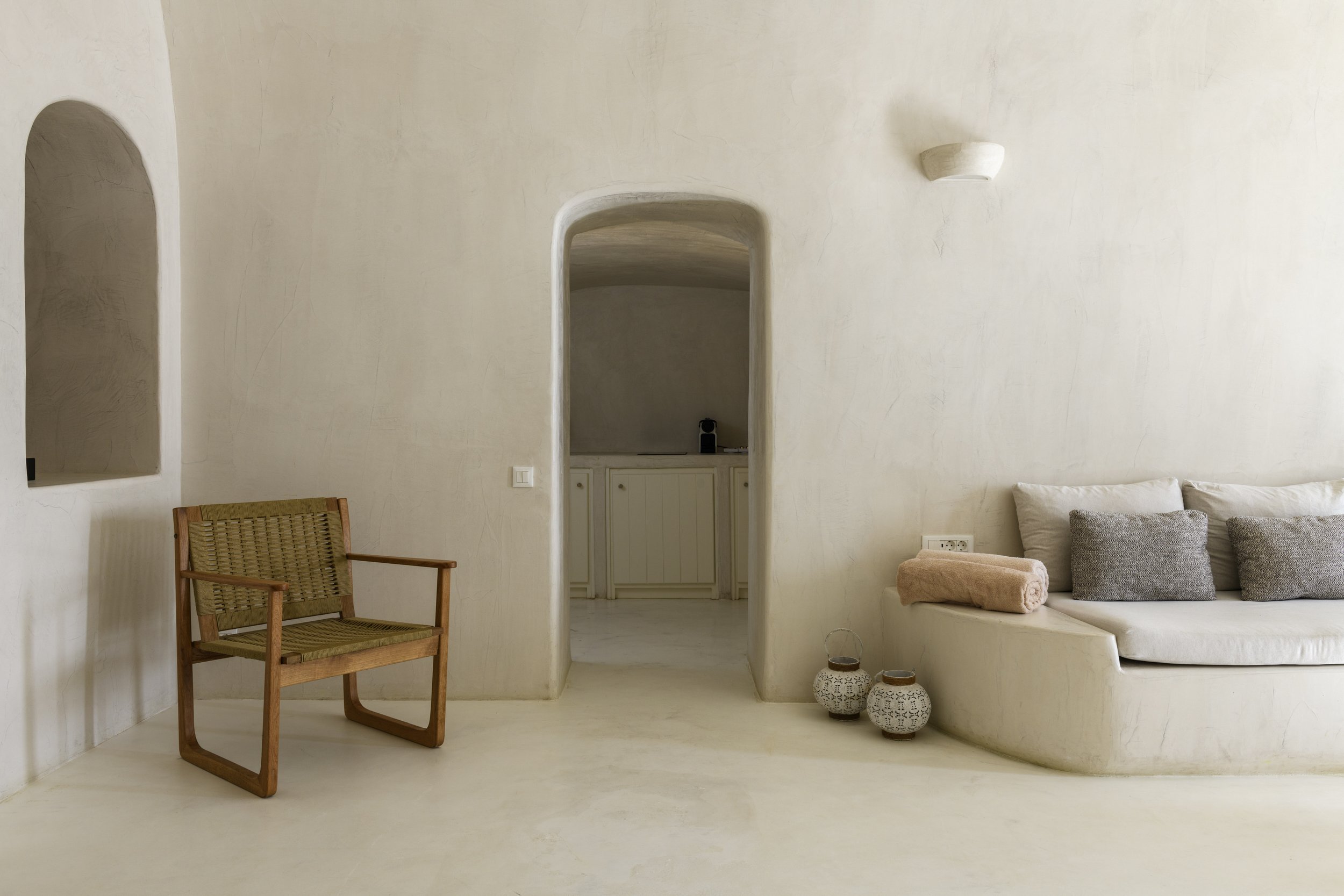 Minimalist living room with adobe-style curved walls, a wicker chair, a built-in curved sofa with pillows, rolled towels, and decorative pottery in neutral tones.