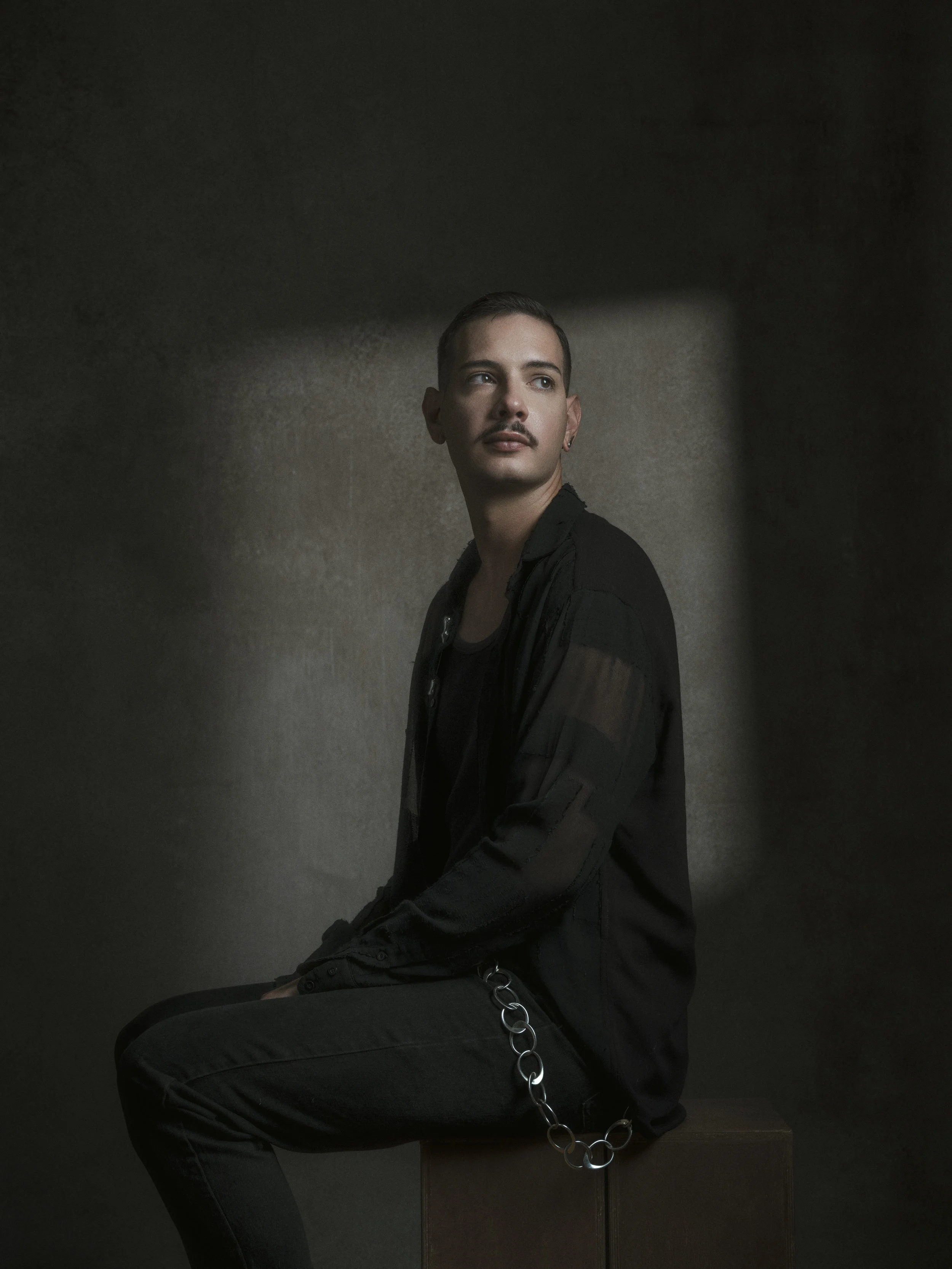 A person wearing black clothing and a chain necklace sitting against a dark wall, with short hairstyle and mustache, looking to the side.