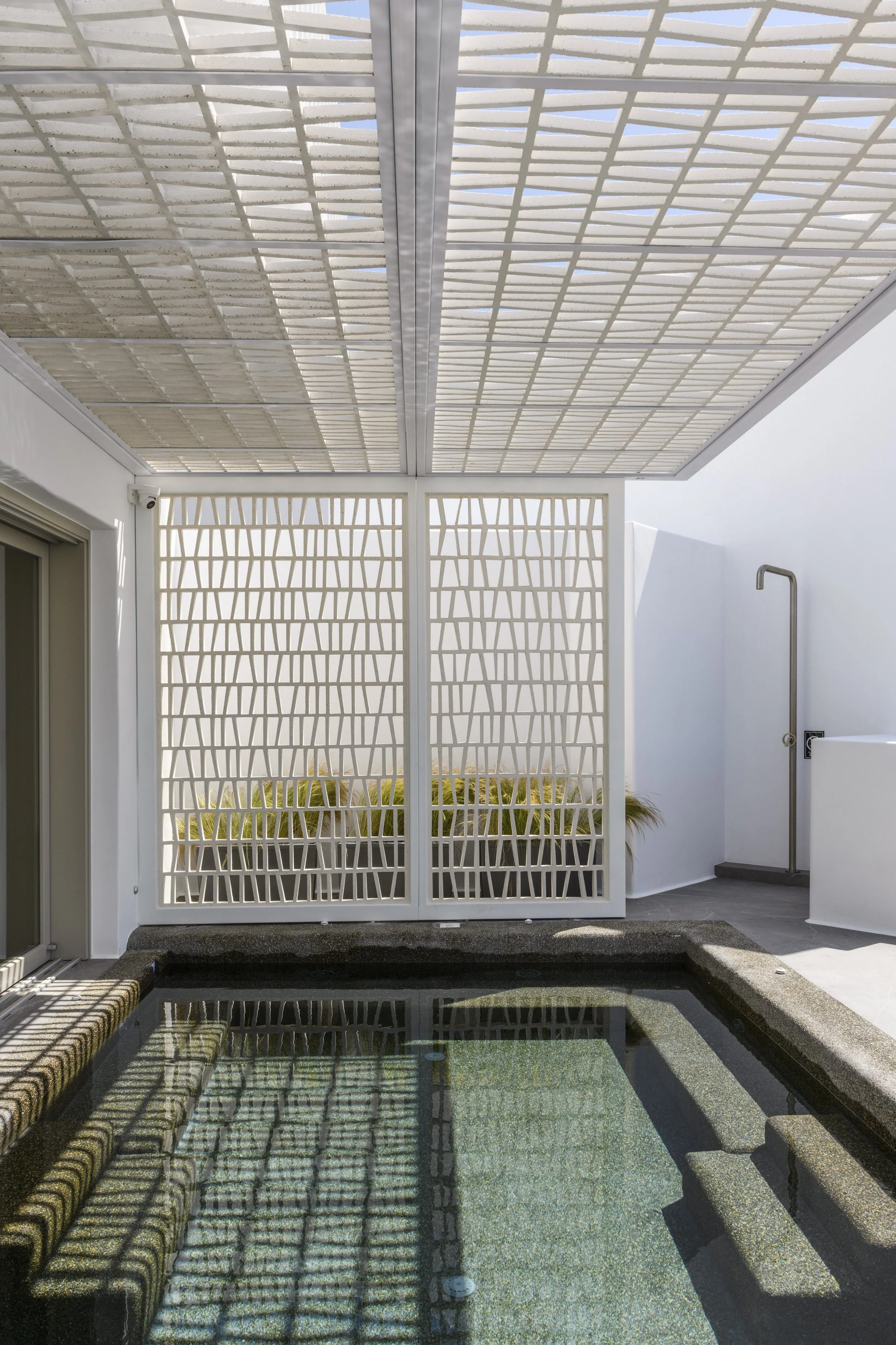 An outdoor space with a small rectangular hot tub or pool, surrounded by a stone or concrete deck. There is a decorative white fence with a geometric pattern, and a white wall with a curved arch. Overhead, a large white pergola or canopy with a slatt
