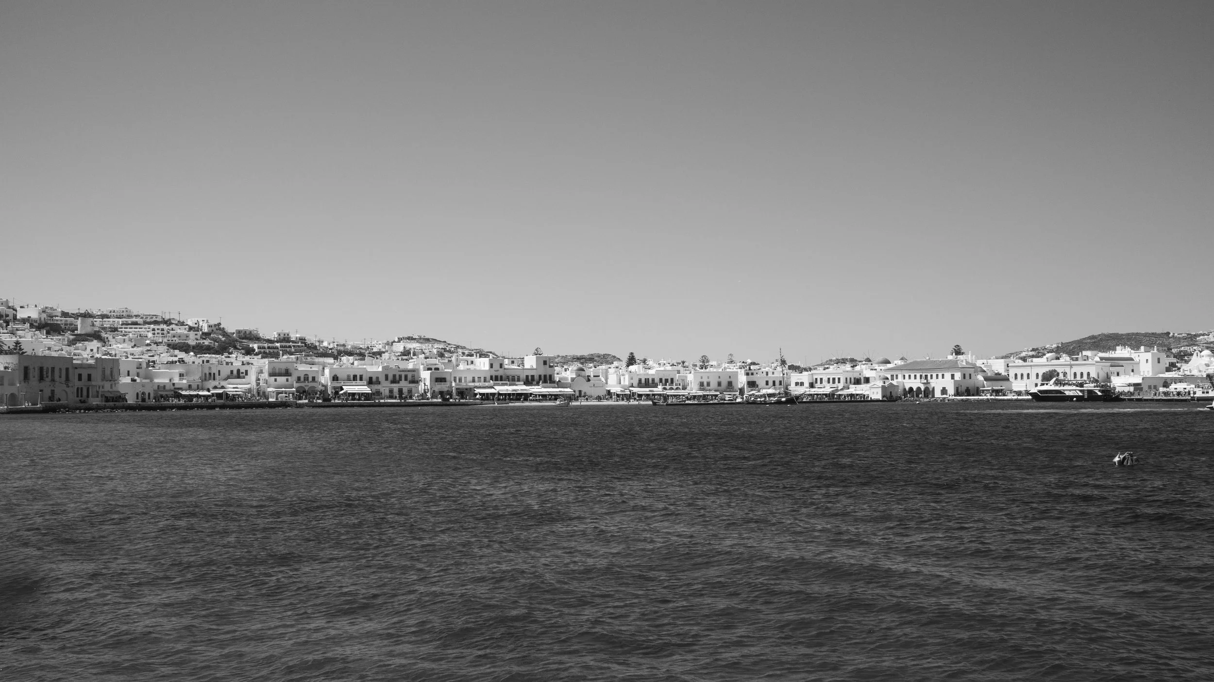 Black and white photo of Mykonos town with white buildings along the shoreline and a boat on the water in the foreground.
Black and white photo of Mykonos town with white buildings along the shoreline and a boat on the water in the foreground.