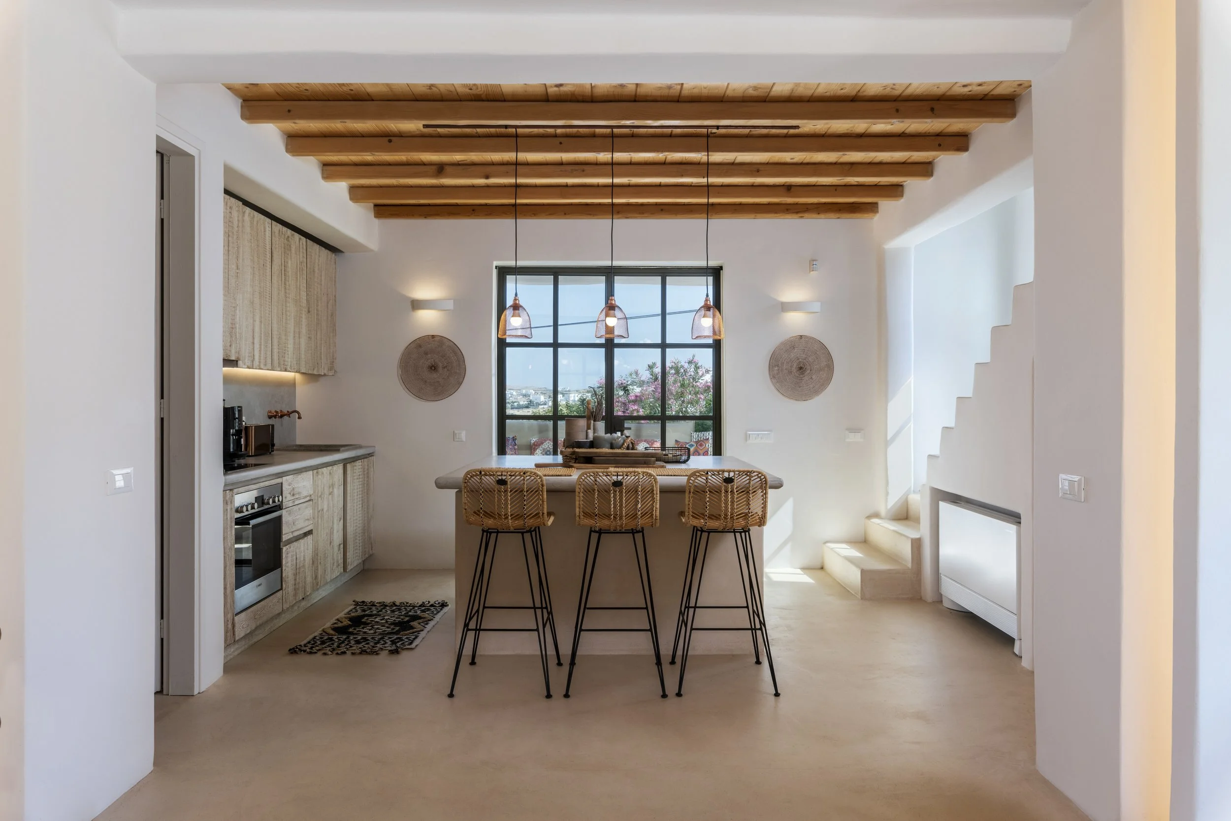 Modern kitchen and dining area with wooden ceiling beams, large window, and high chairs at a breakfast bar.