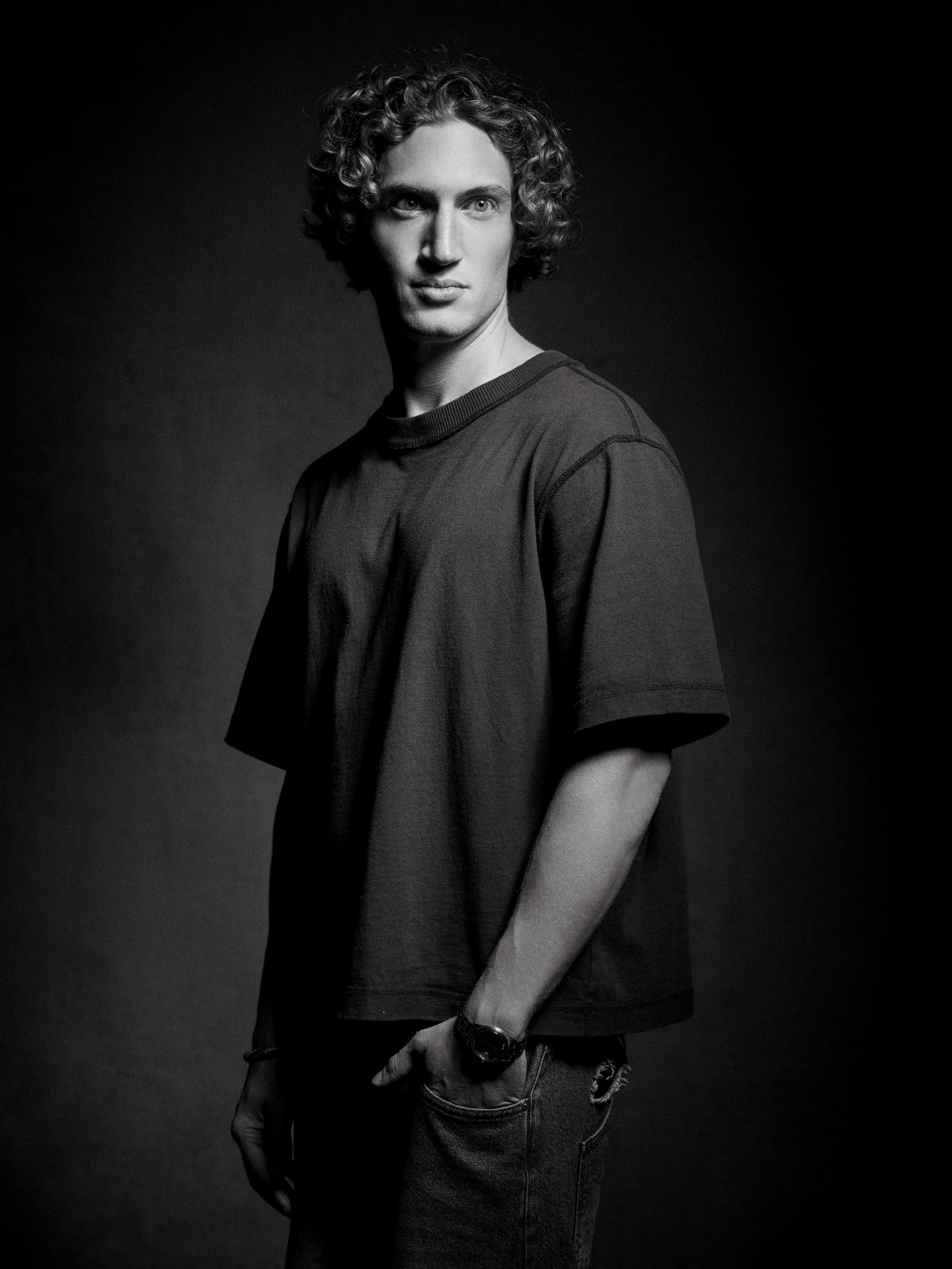 Black and white photo of a young person with curly hair, wearing a short-sleeved T-shirt, denim shorts, and a watch, standing against a dark background.