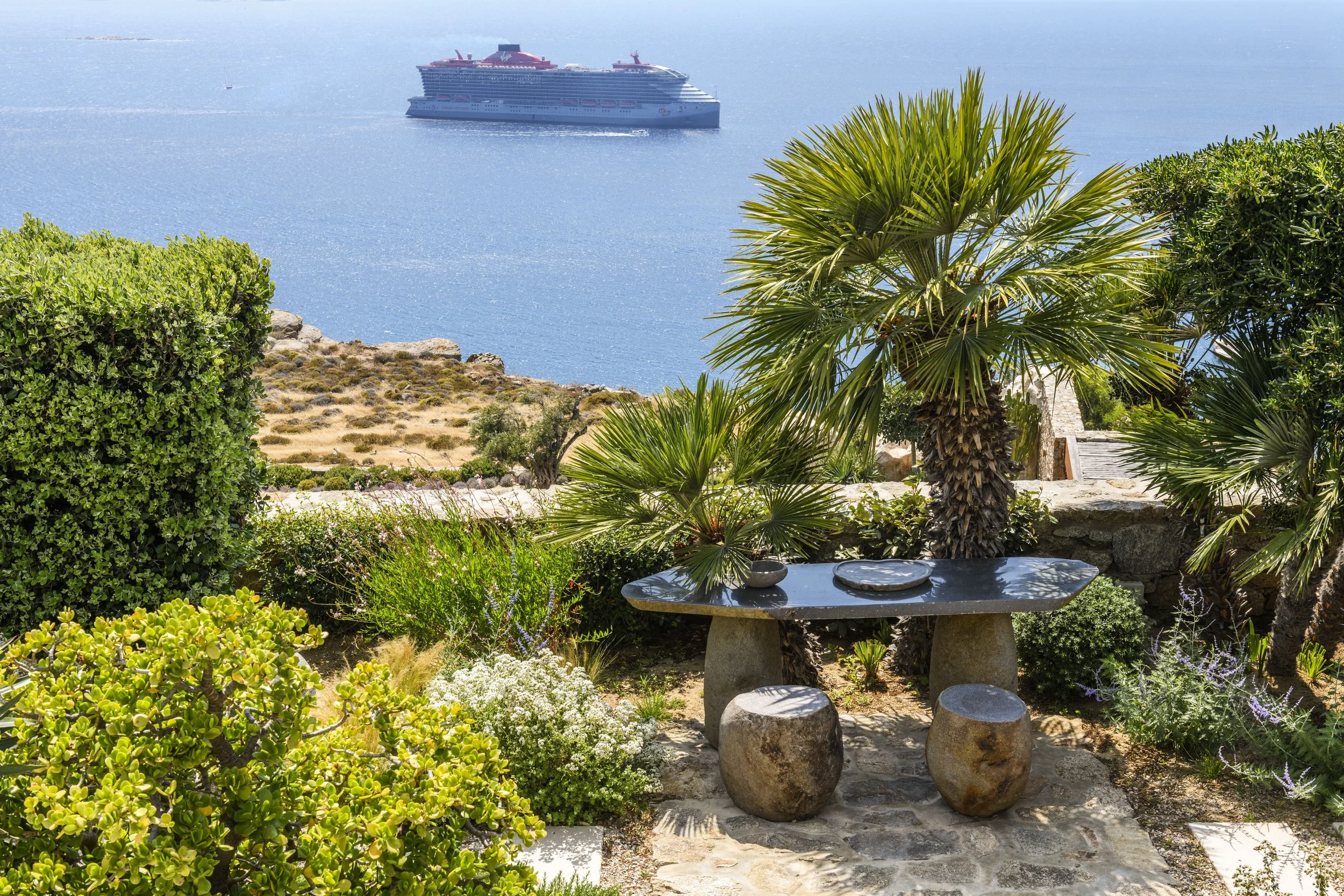 Outdoor stone terrace and seating area surrounded by Mediterranean greenery at a luxury Mykonos island estate, overlooking a cruise ship on the deep blue sea.