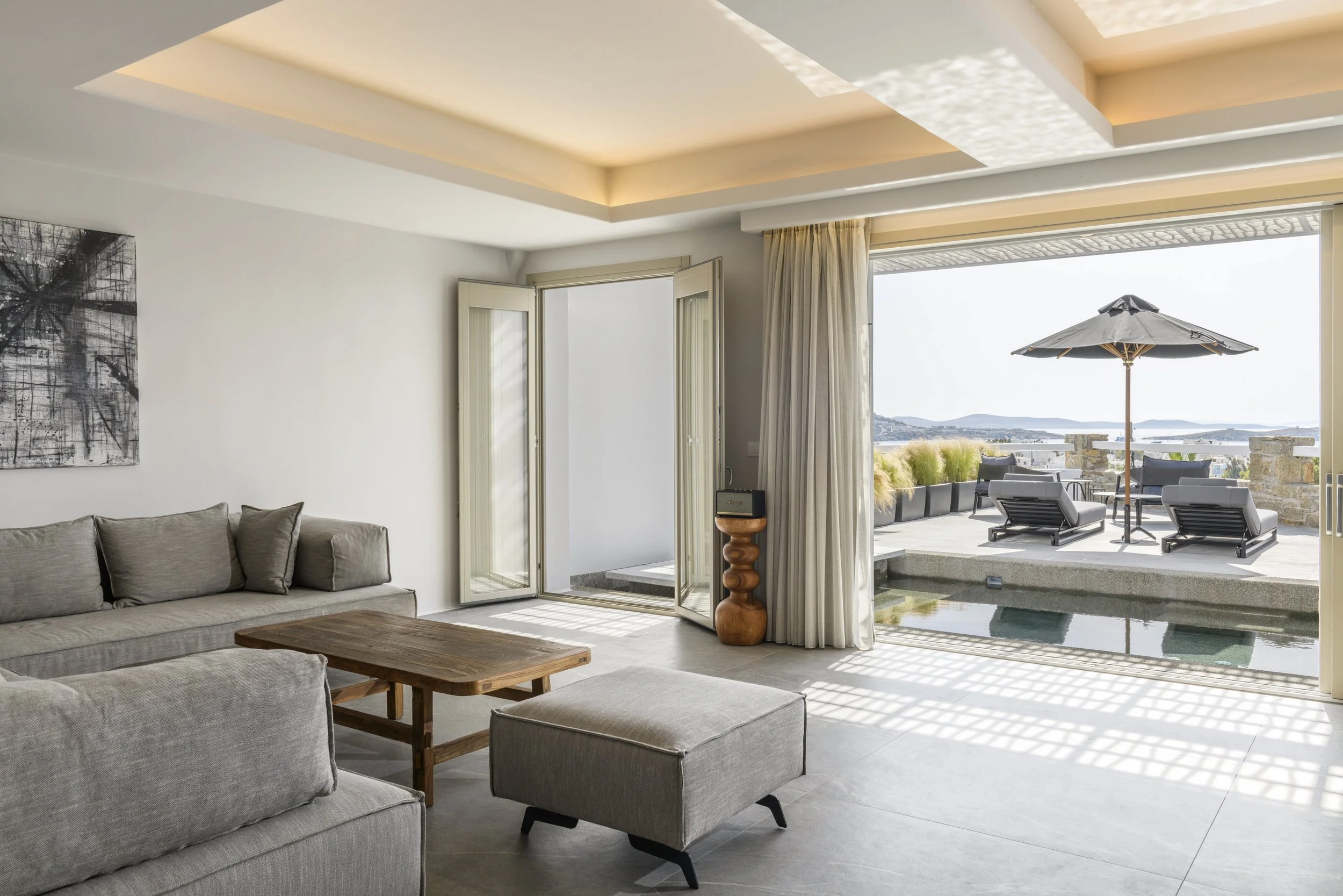 Living room with beige sofas, wooden coffee table, open balcony door leading to outdoor seating area with umbrella, view of water and sky outside.