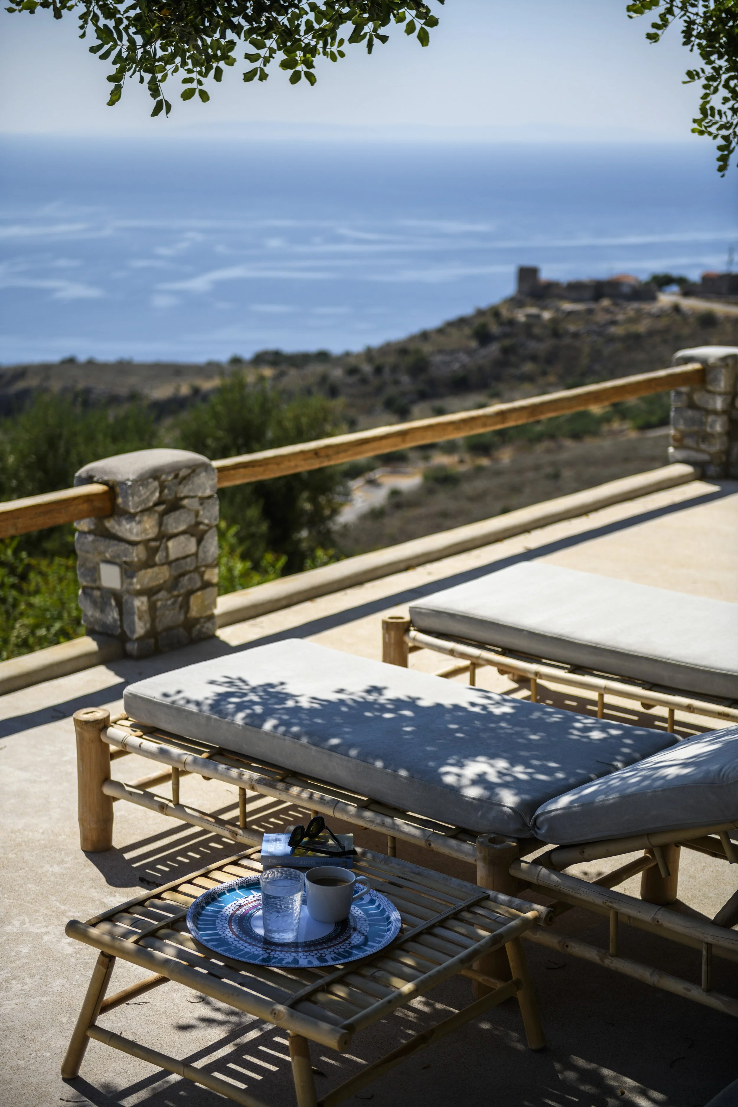Outdoor patio with bamboo lounge chairs and a small table set with drinks and cups, overlooking a hilly landscape with an ancient castle in the distance, under a tree with green leaves.