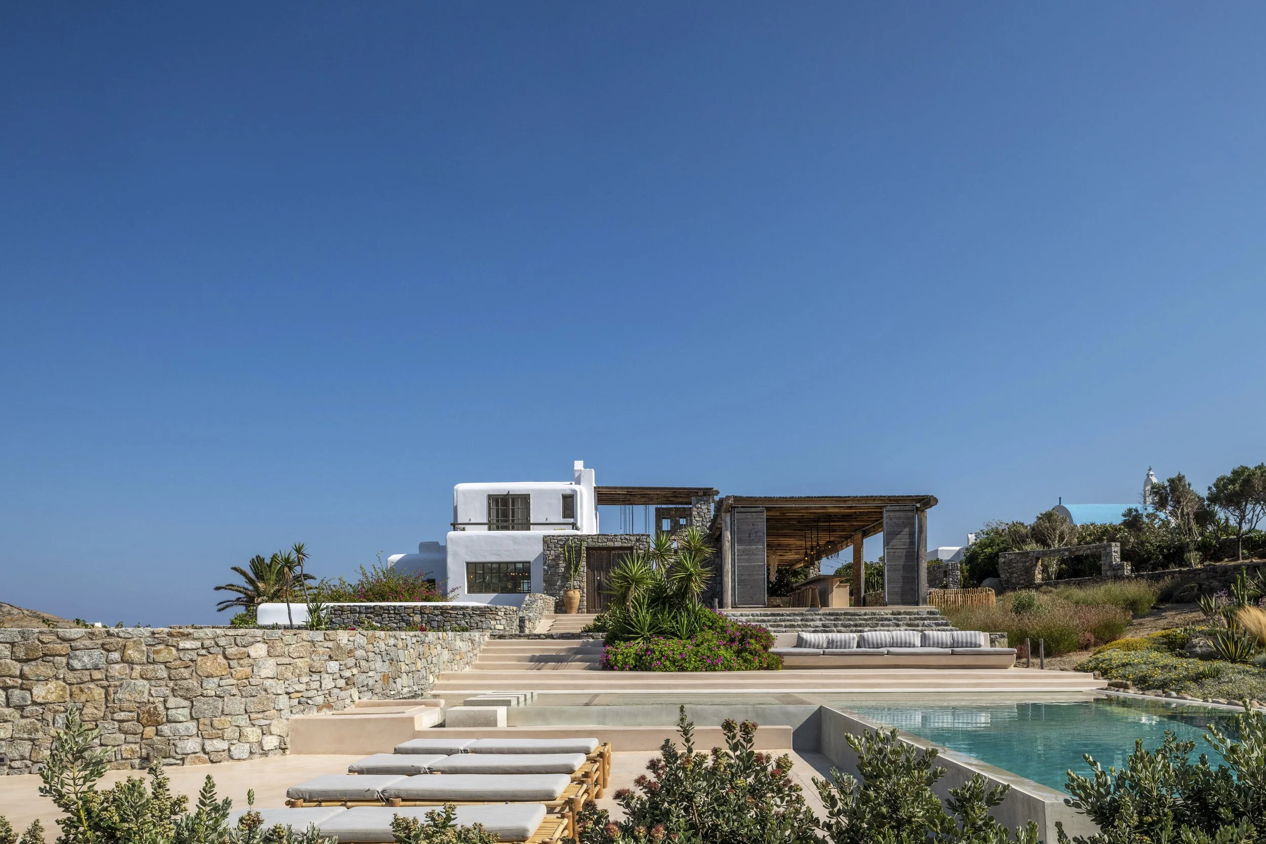 Modern white house with flat roof and large windows, overlooking a swimming pool and landscaped garden with plants and stairs, under a blue sky.