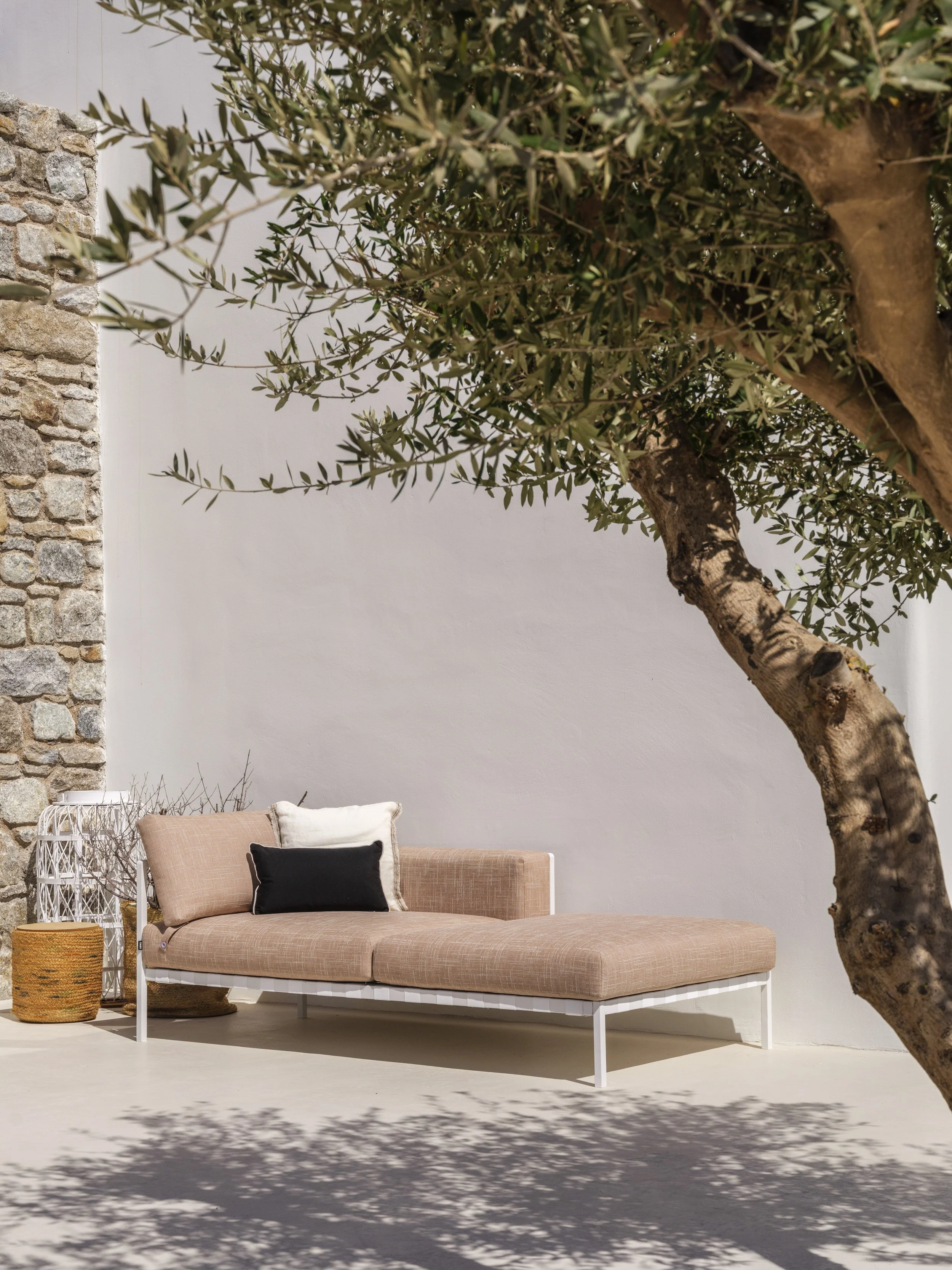 A beige cushioned outdoor sofa with white metal frame, accented with a black pillow and a white pillow, set against a plain white wall next to a stone wall, with a large tree casting shadows in the foreground.