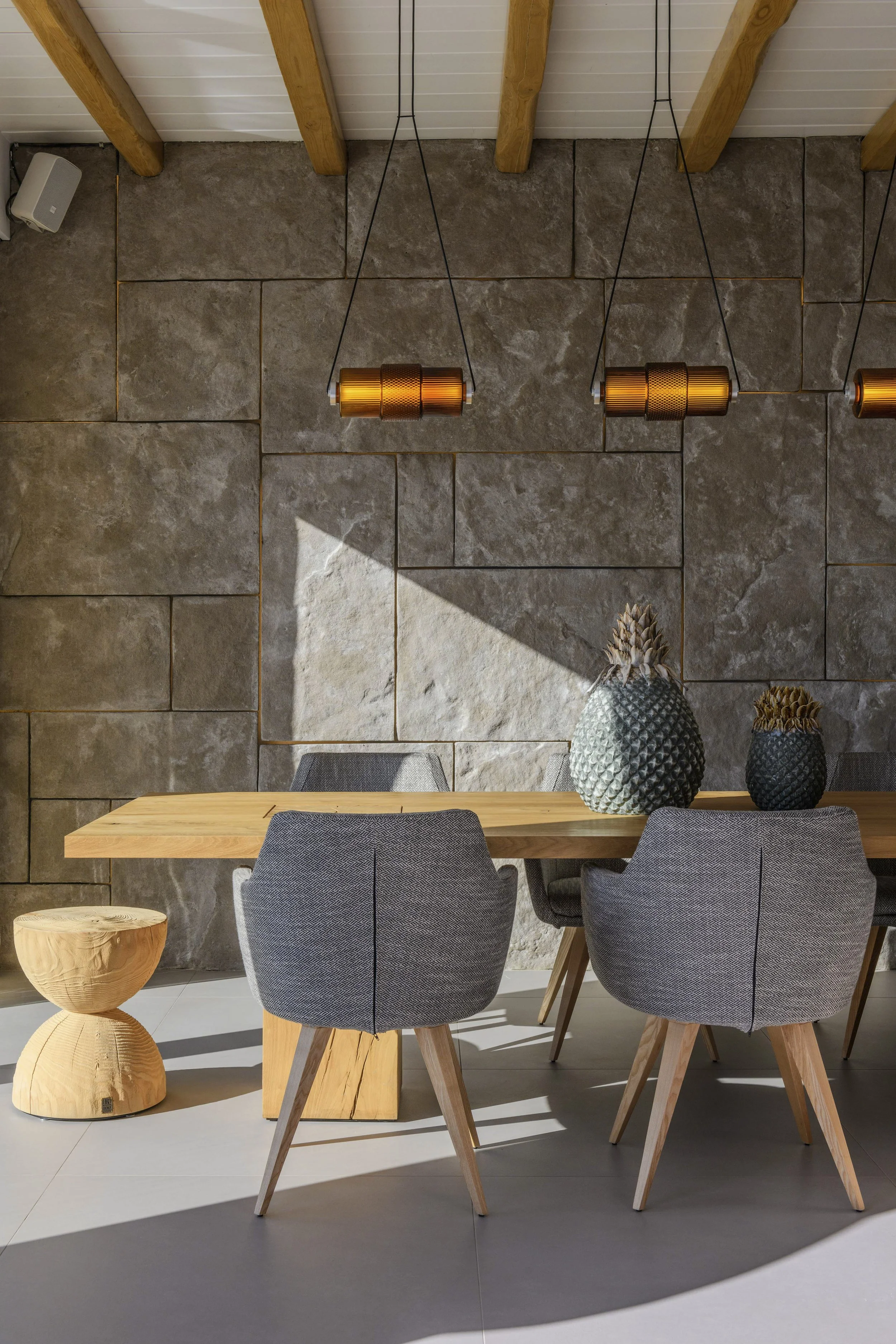A modern dining area featuring a wooden table, four gray upholstered chairs with wooden legs, a small wooden side table, and decorative vases on the table, with a textured stone accent wall and hanging copper pendant lights.
