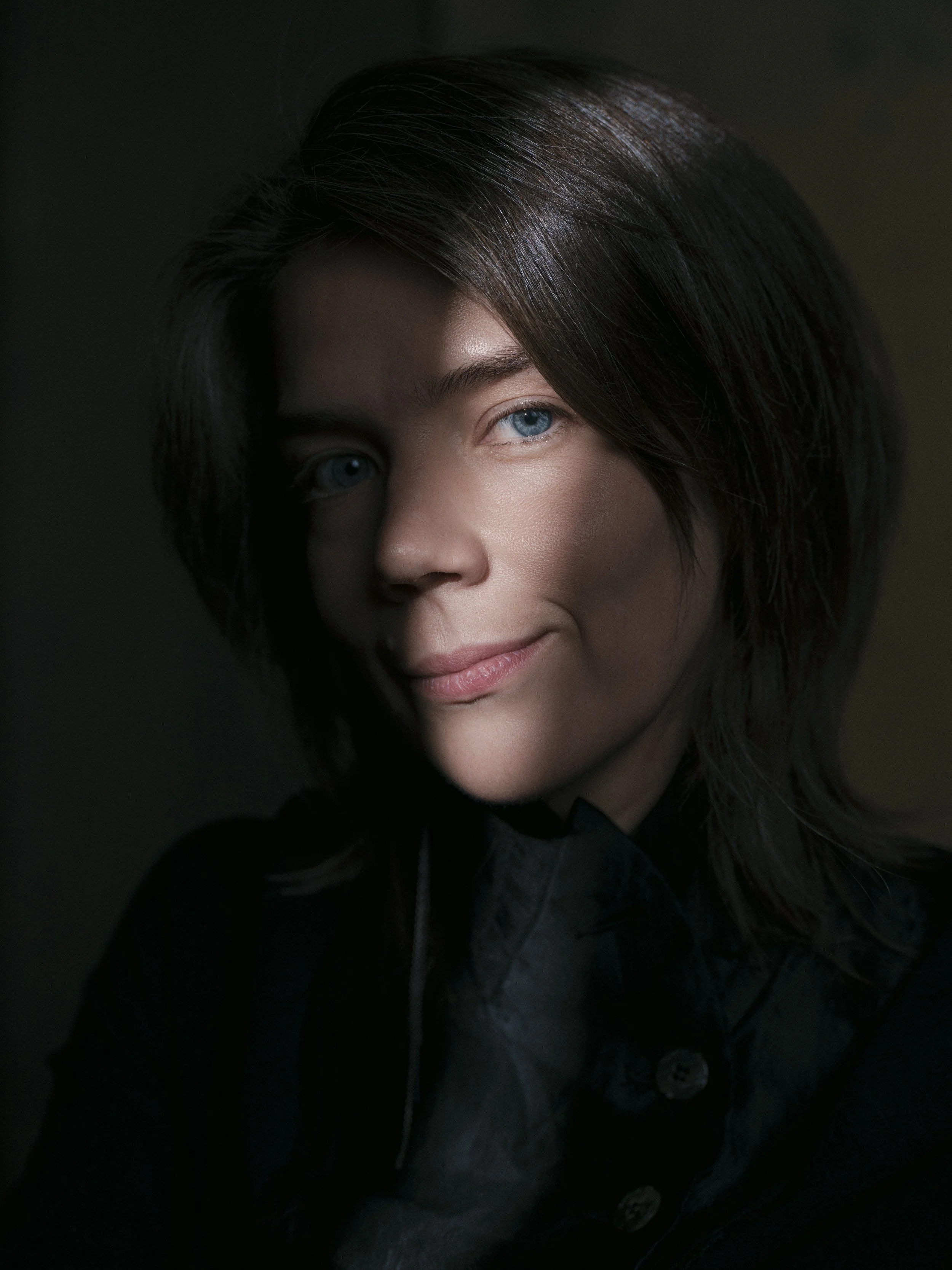 Close-up of a woman with dark brown hair, blue eyes, and light skin, smiling softly, with shadows cast on her face.