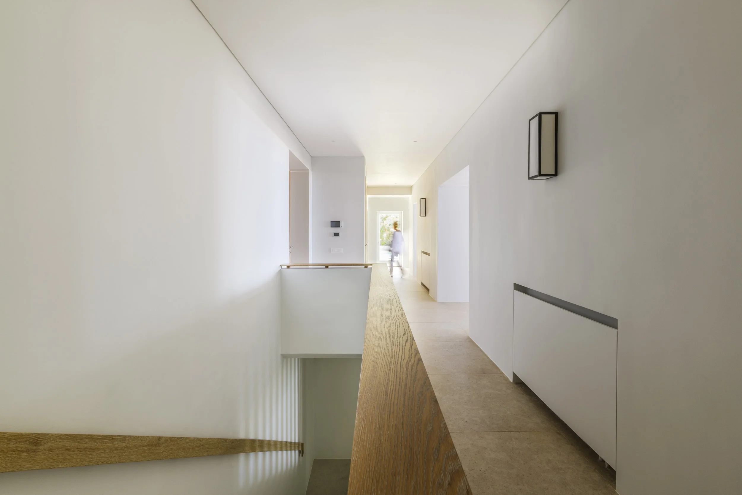 A modern, minimalist hallway with white walls and ceiling, natural light from a window, a wood banister, a wall-mounted light fixture, and a woman in the background walking near the open door.