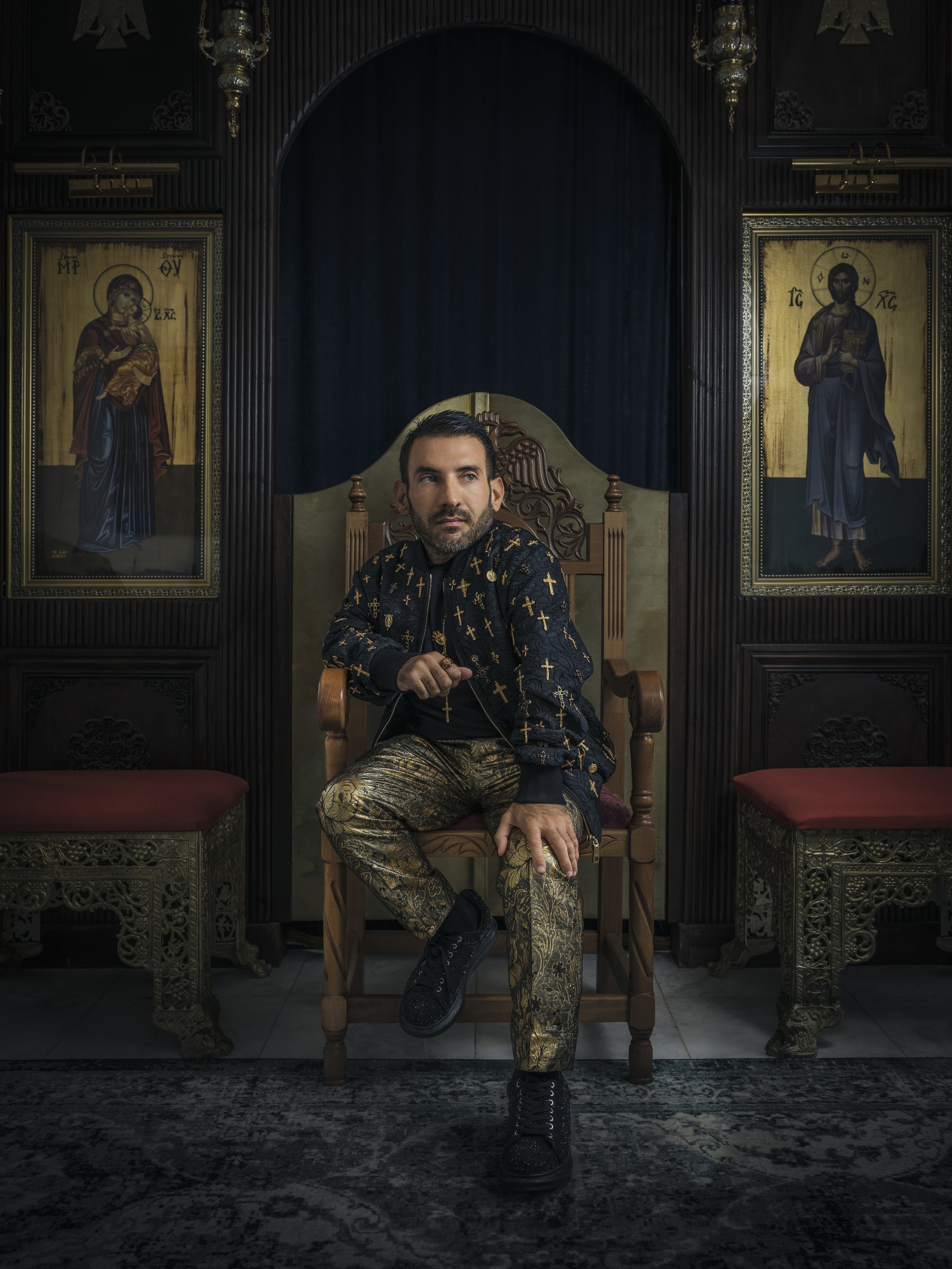 A man sitting on a wooden throne-like chair in a dark, ornate room with religious icons on the wall behind him. He is wearing a black jacket with gold crosses, gold patterned pants, and black shoes.