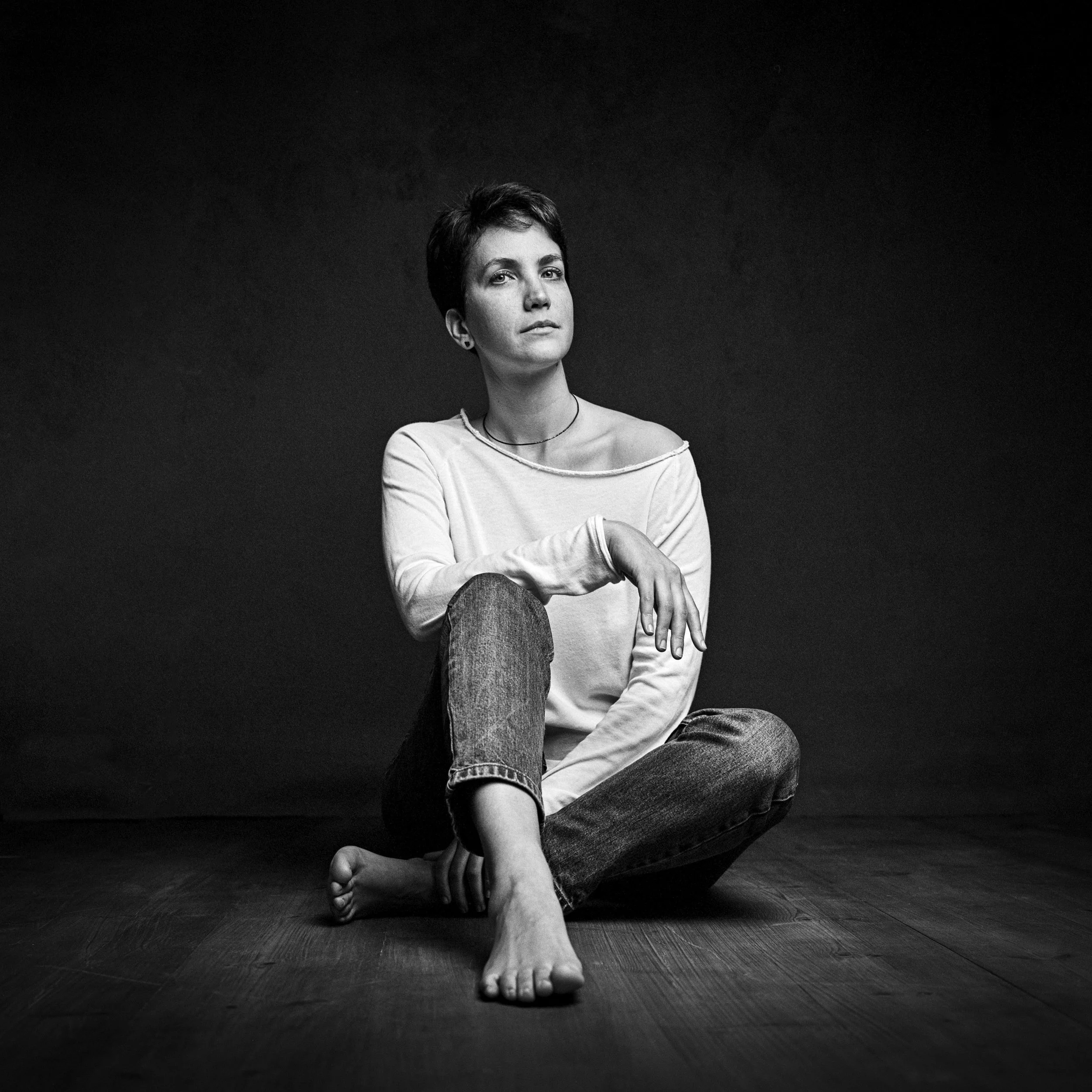 Black and white photo of a woman with short hair, sitting on a hardwood floor against a dark background, in a relaxed pose with one arm resting on her knee and the other on her ankle.