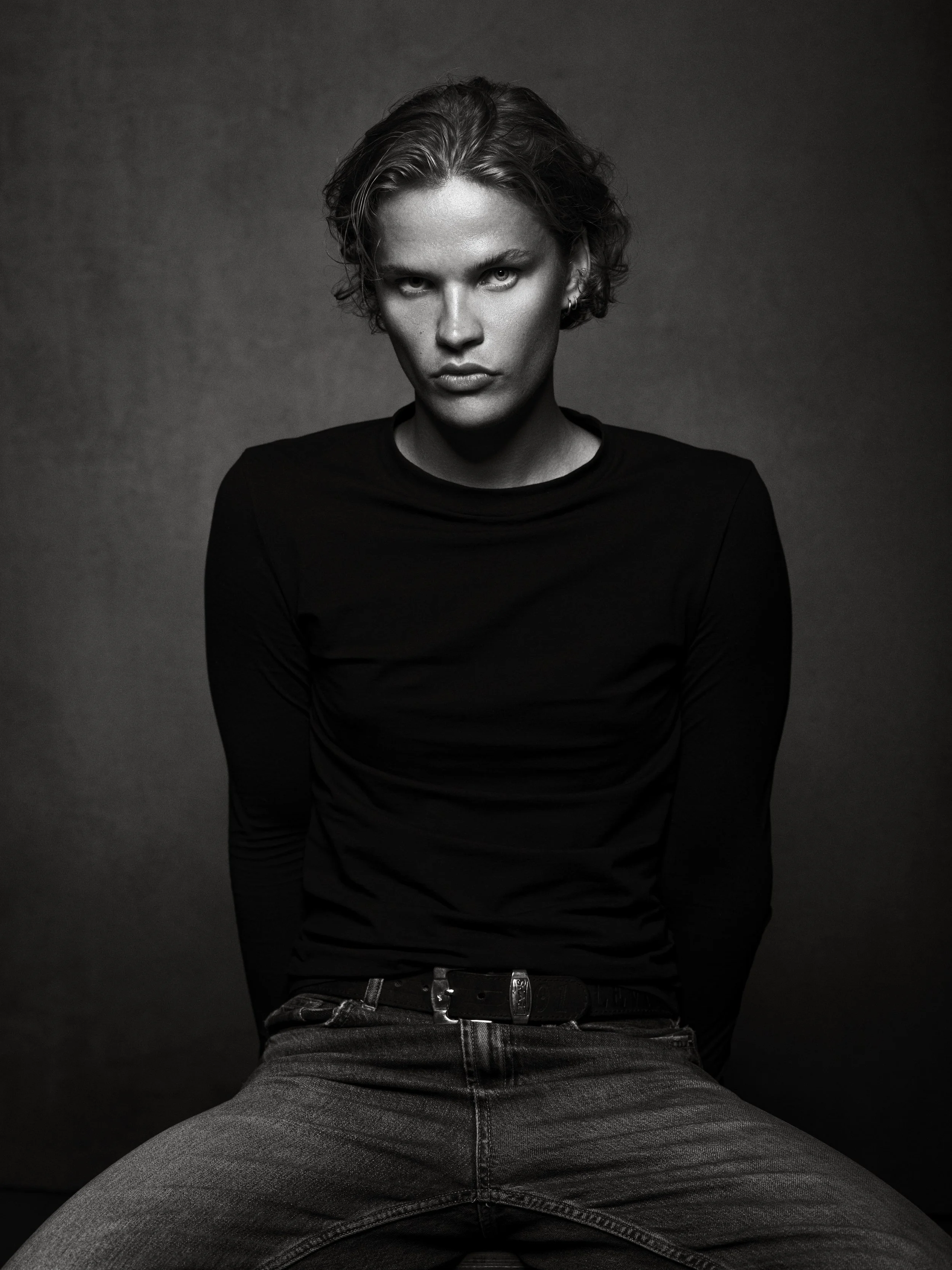 Black and white photo of a woman with short wavy hair, wearing a black long-sleeve shirt and jeans, sitting with her legs apart and looking directly at the camera.