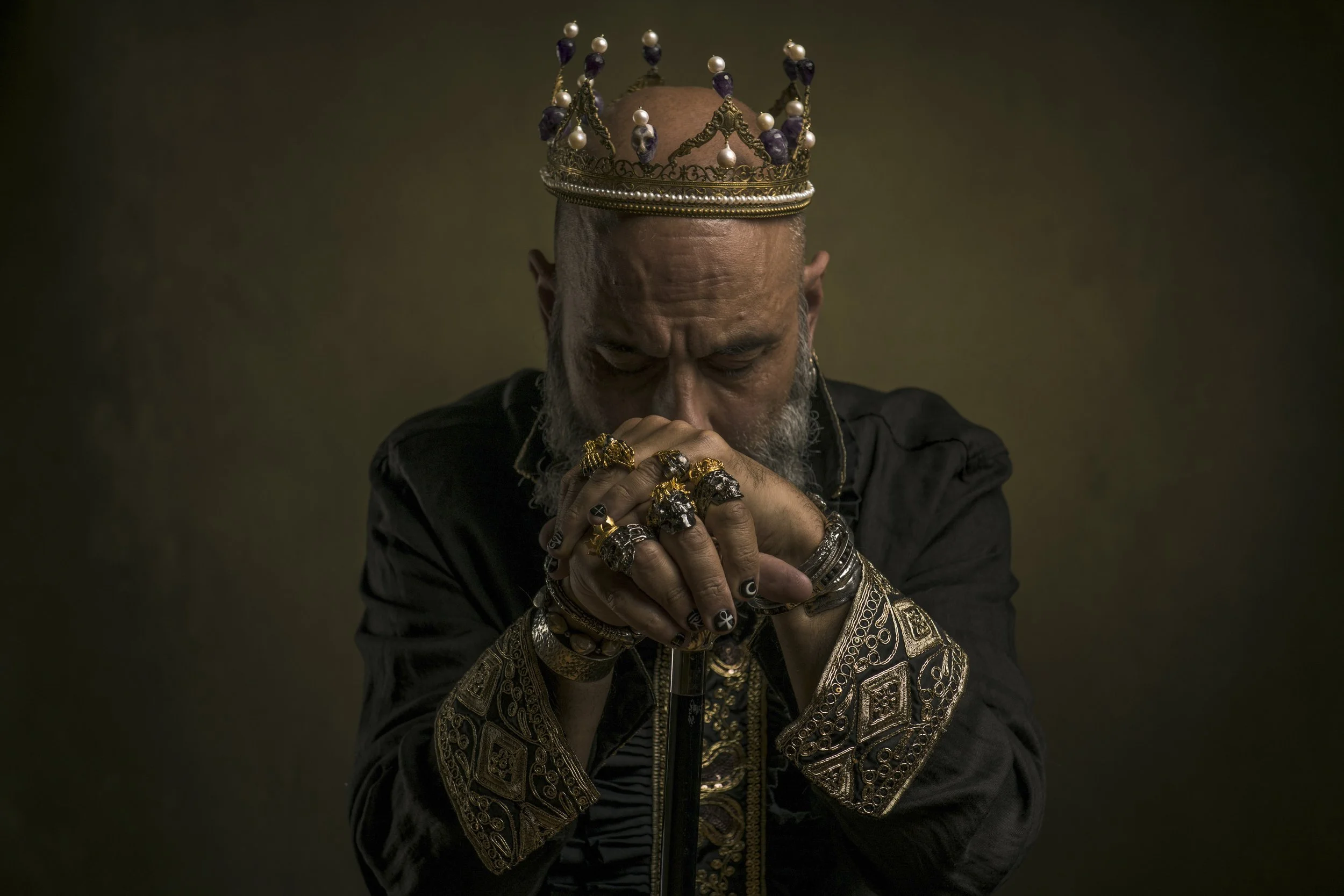 A man with a shaved head and beard wearing numerous gold rings, bracelets, and rings on his fingers, wearing a black jacket with gold embroidery, with his eyes closed and hands clasped in front of his face. He is also wearing a gold crown decorated w