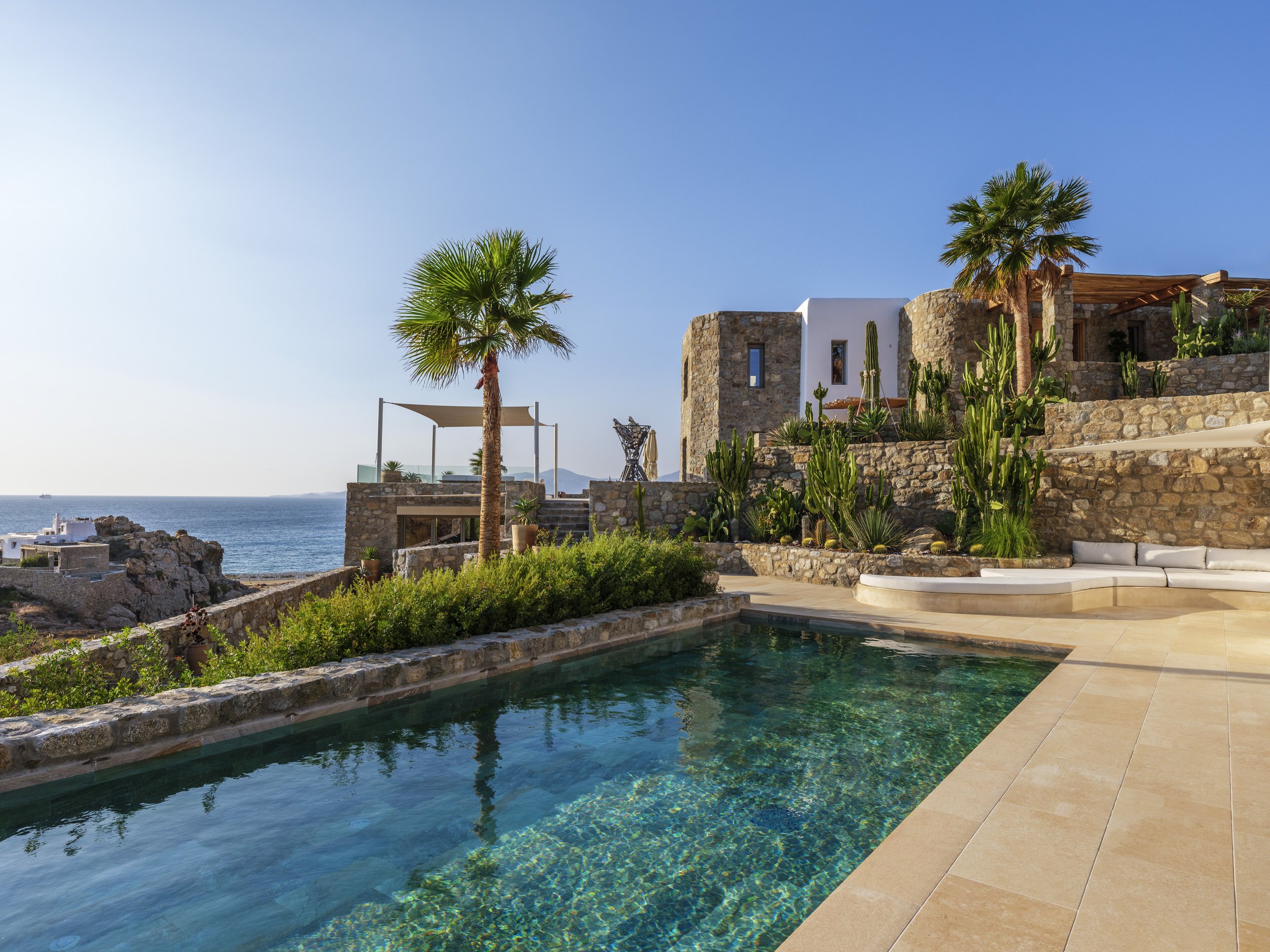 An outdoor swimming pool with arched stone building, palm trees, and cactus plants overlooking the ocean under a clear blue sky.