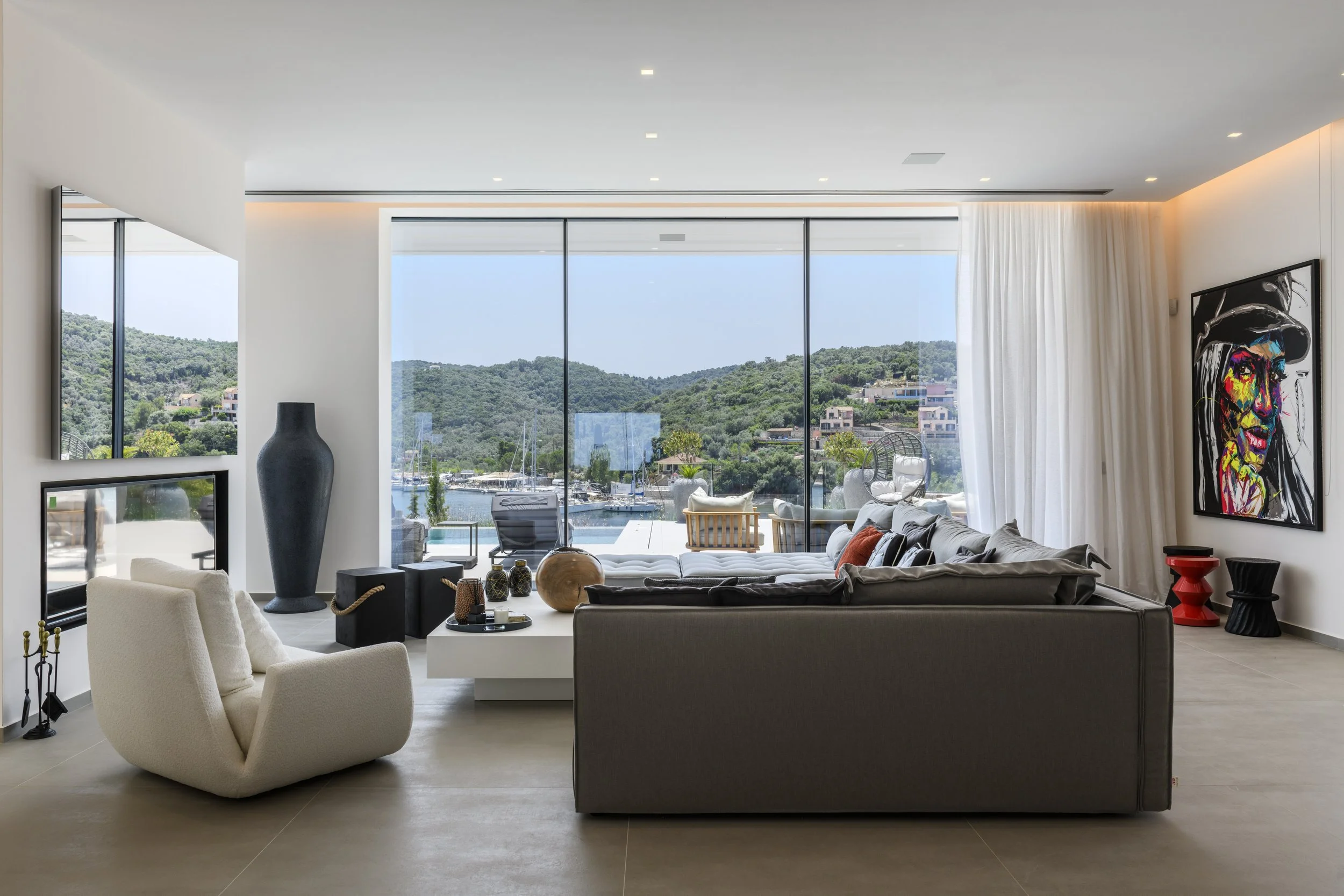 Modern living room with large glass windows overlooking a hillside and marina, featuring white and gray furniture, colorful wall art, and decorative objects.