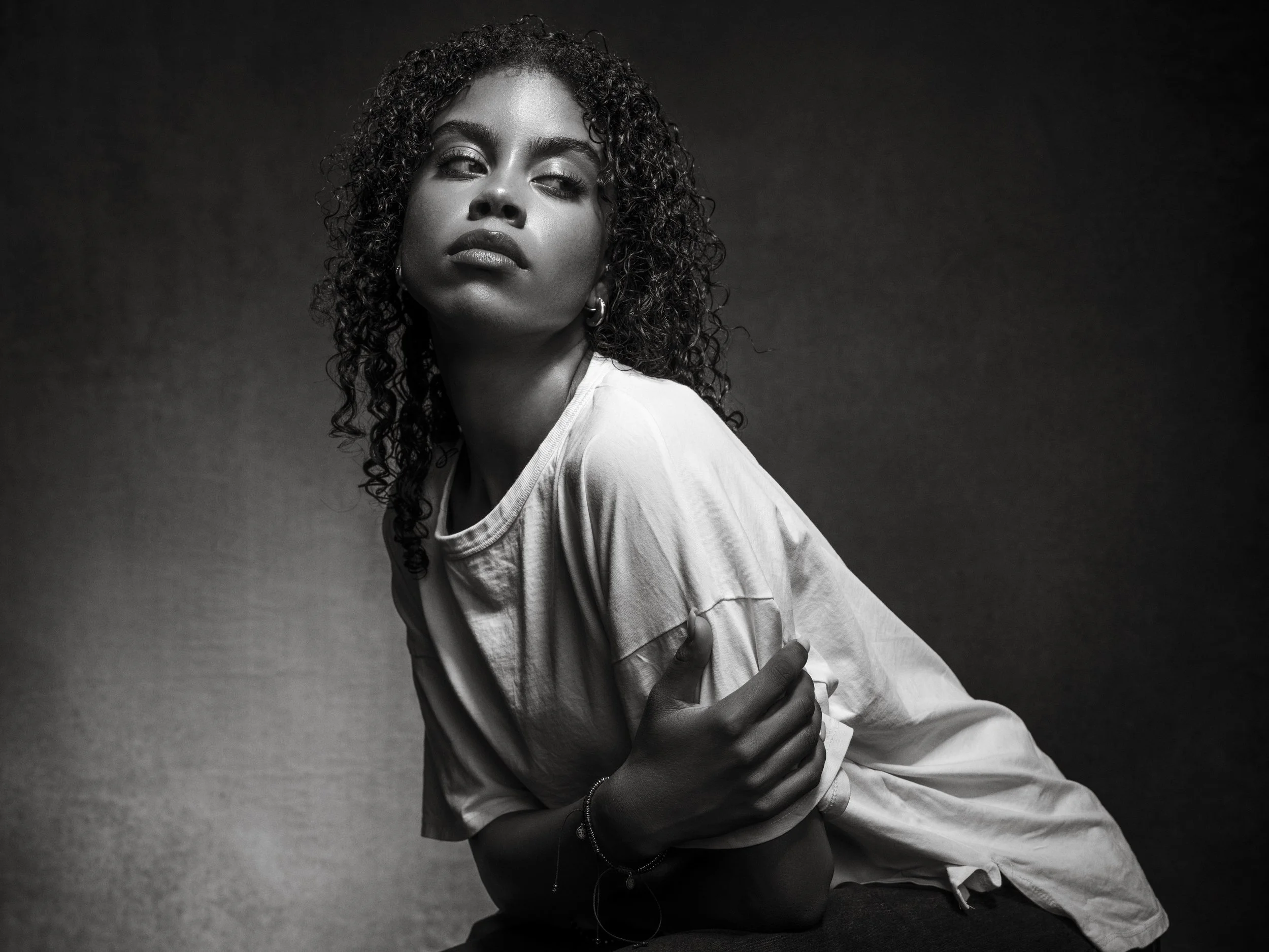 Black and white portrait of a woman with curly hair, looking to the side with a serious expression, wearing a loose shirt and jewelry against a dark background.