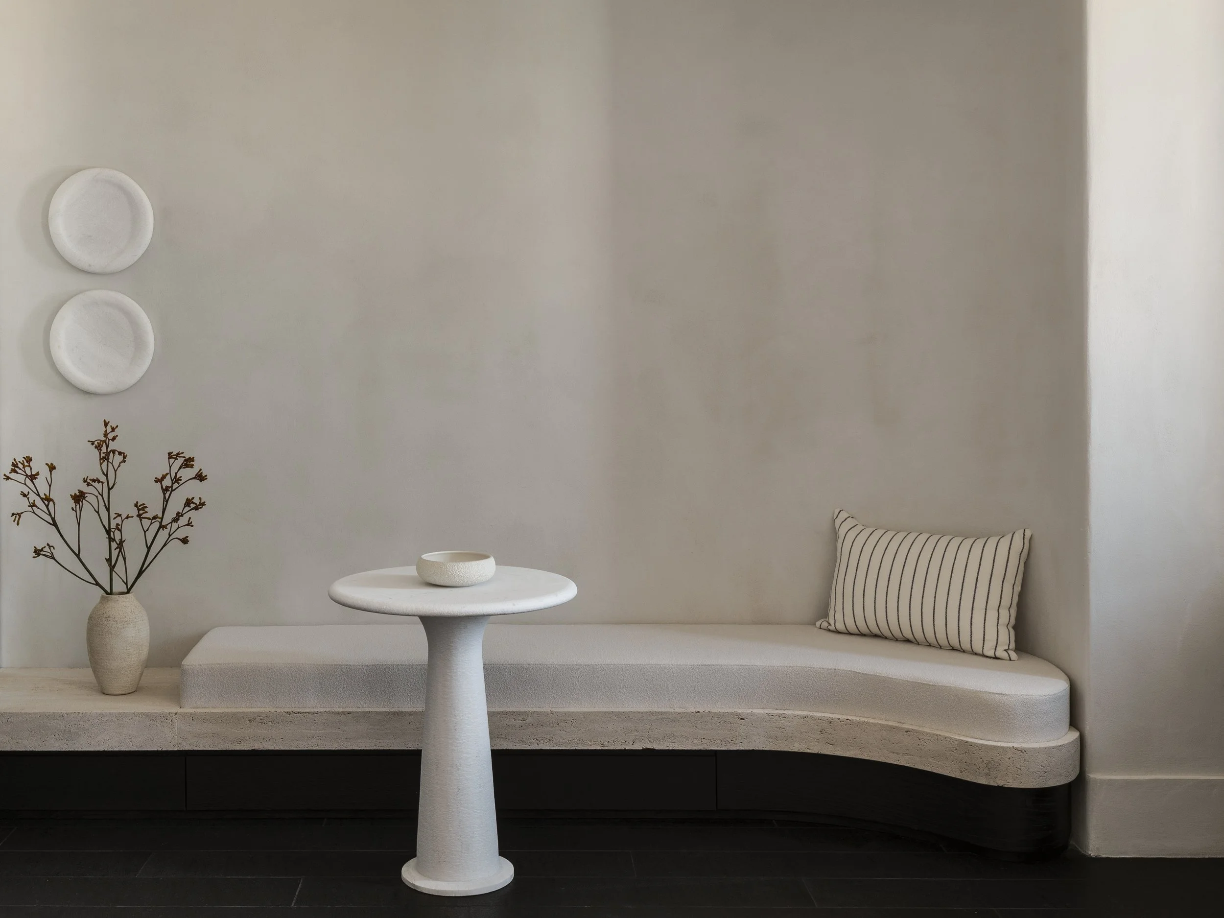 A minimalist interior scene with a white curved bench built into a wall, a white round side table, a white ceramic bowl, a large beige vase with dried branches, and a striped pillow on the bench. The wall is plain and light-colored, creating a calm and neutral atmosphere.
