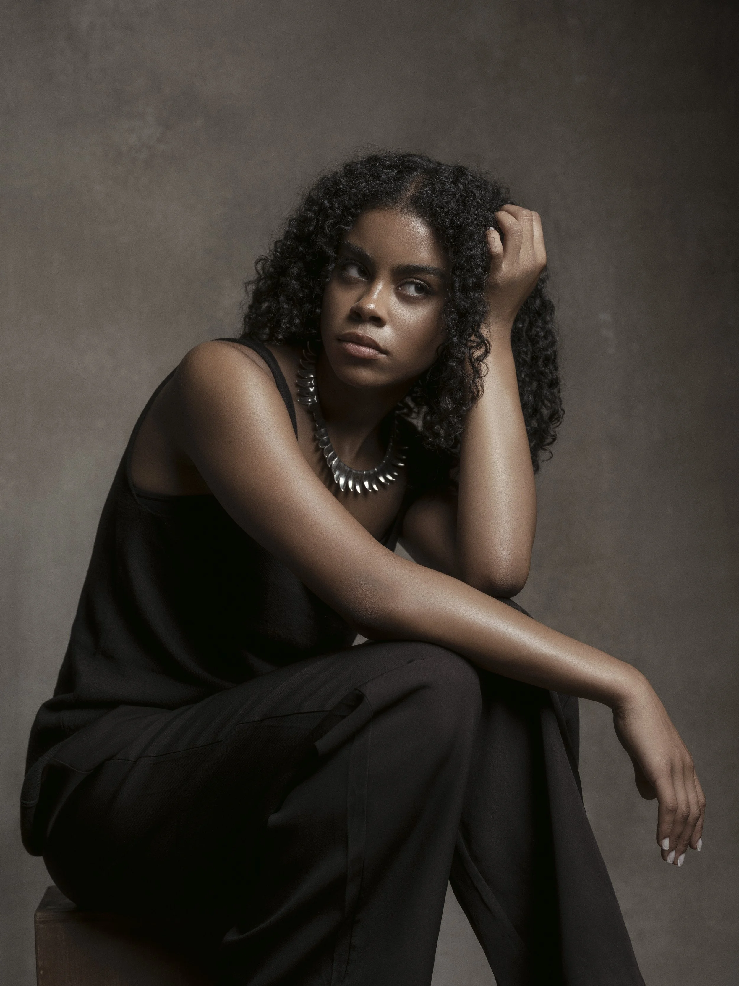 A young woman with dark, curly hair sitting on a wooden block, wearing a black sleeveless top and a chunky metallic necklace, against a plain gray background.
