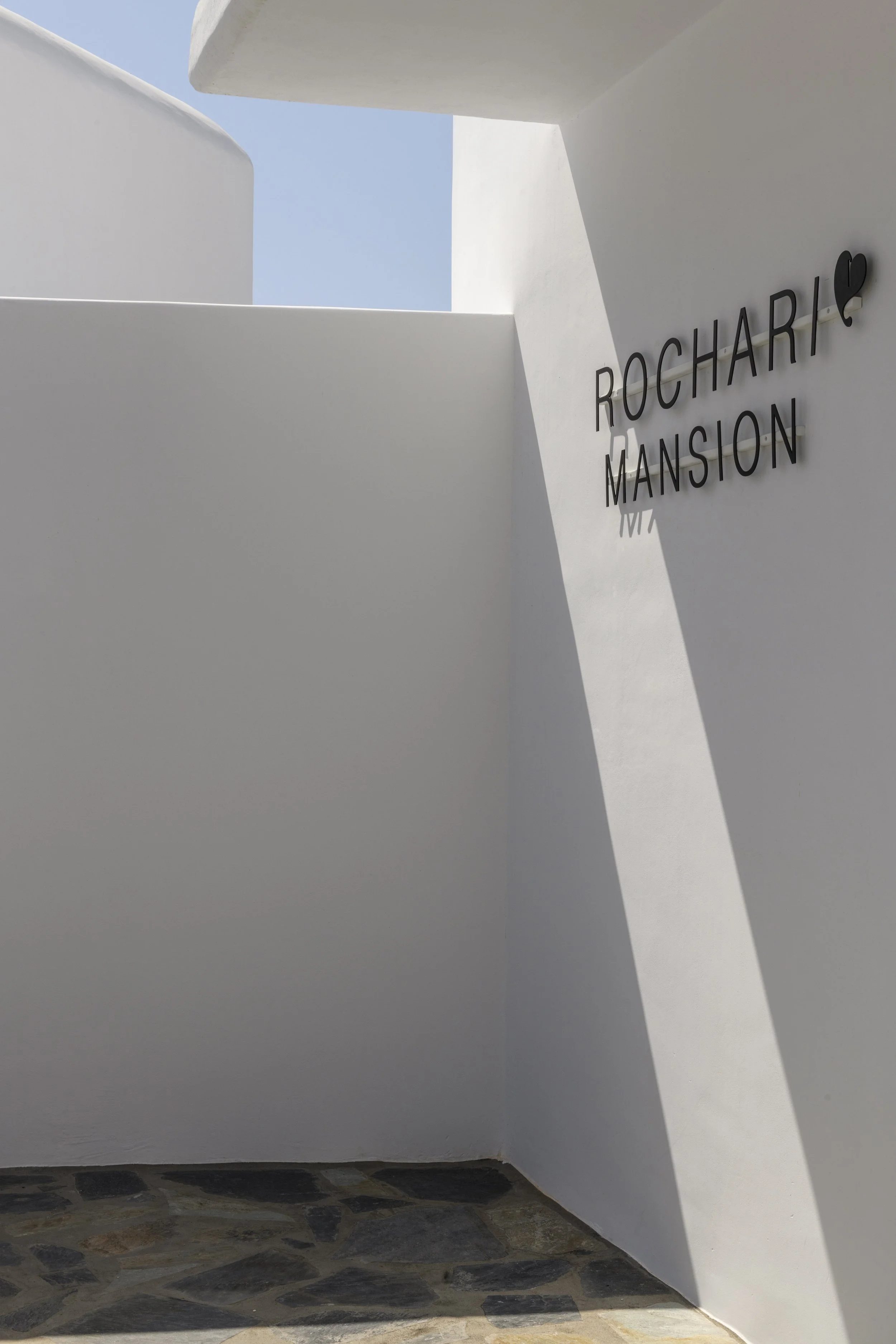 White modern building with simple design, the sign 'Rocharc Mansion' with a heart symbol, and a blue sky visible above.