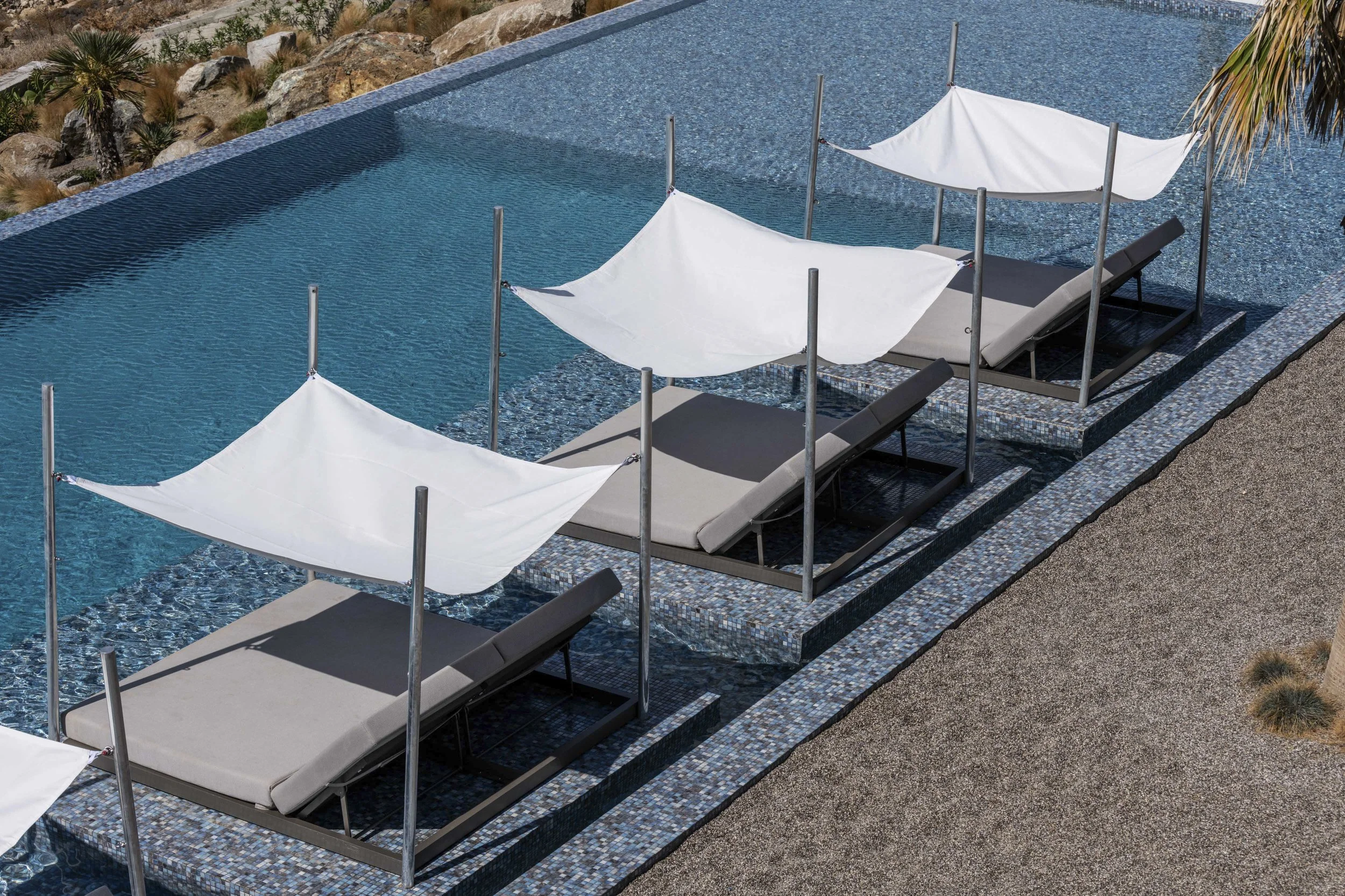 Swimming pool with three lounge chairs and white shade sails, surrounded by desert rocks and plants, pool with mosaic tile edge overlooking the Aegean sea and distant islands under a clear blue sky located in Mykonos island Greece
