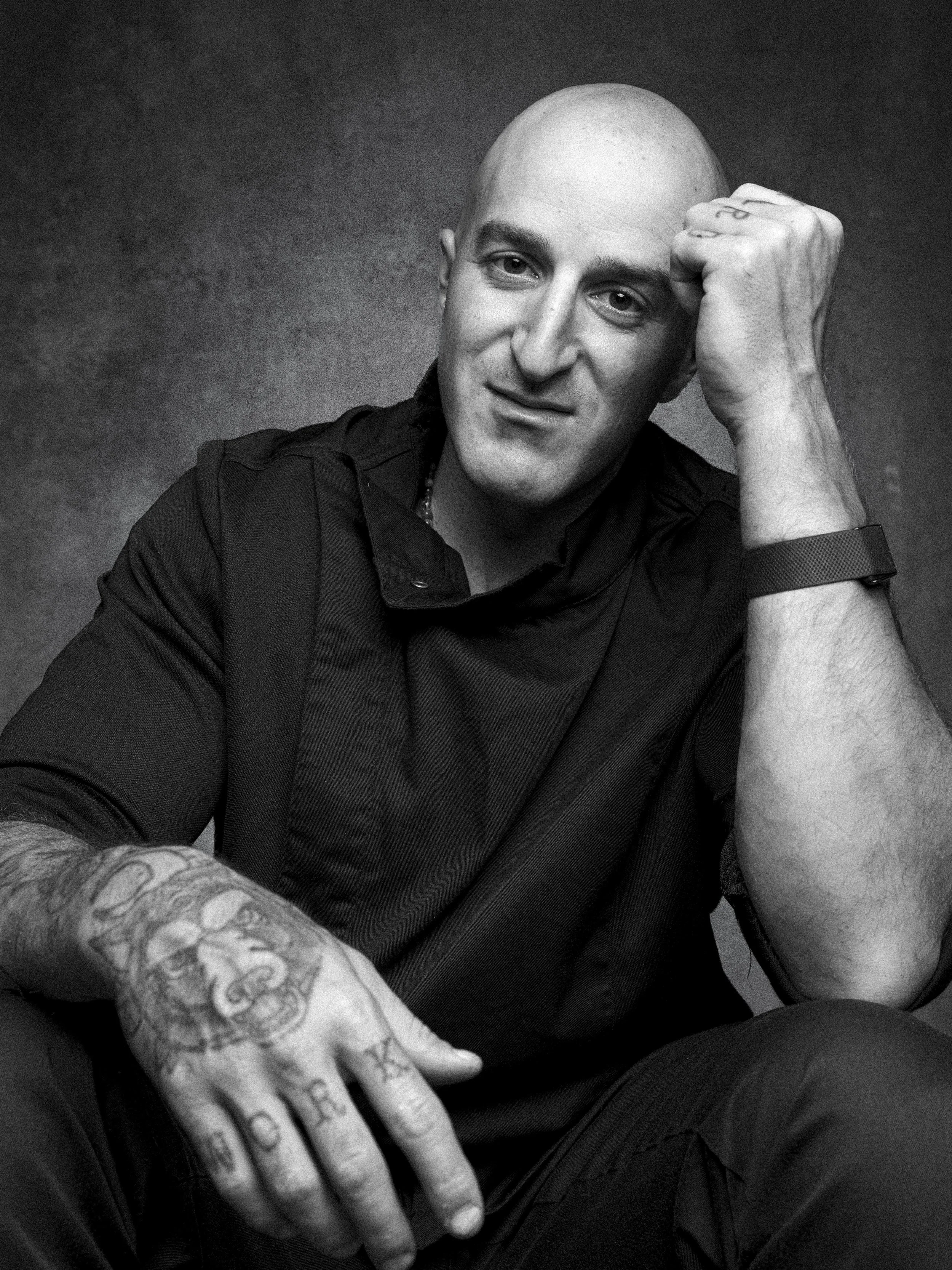Black and white portrait of a bald man with a tattooed hand resting on his knee, looking at the camera with a slight smile.