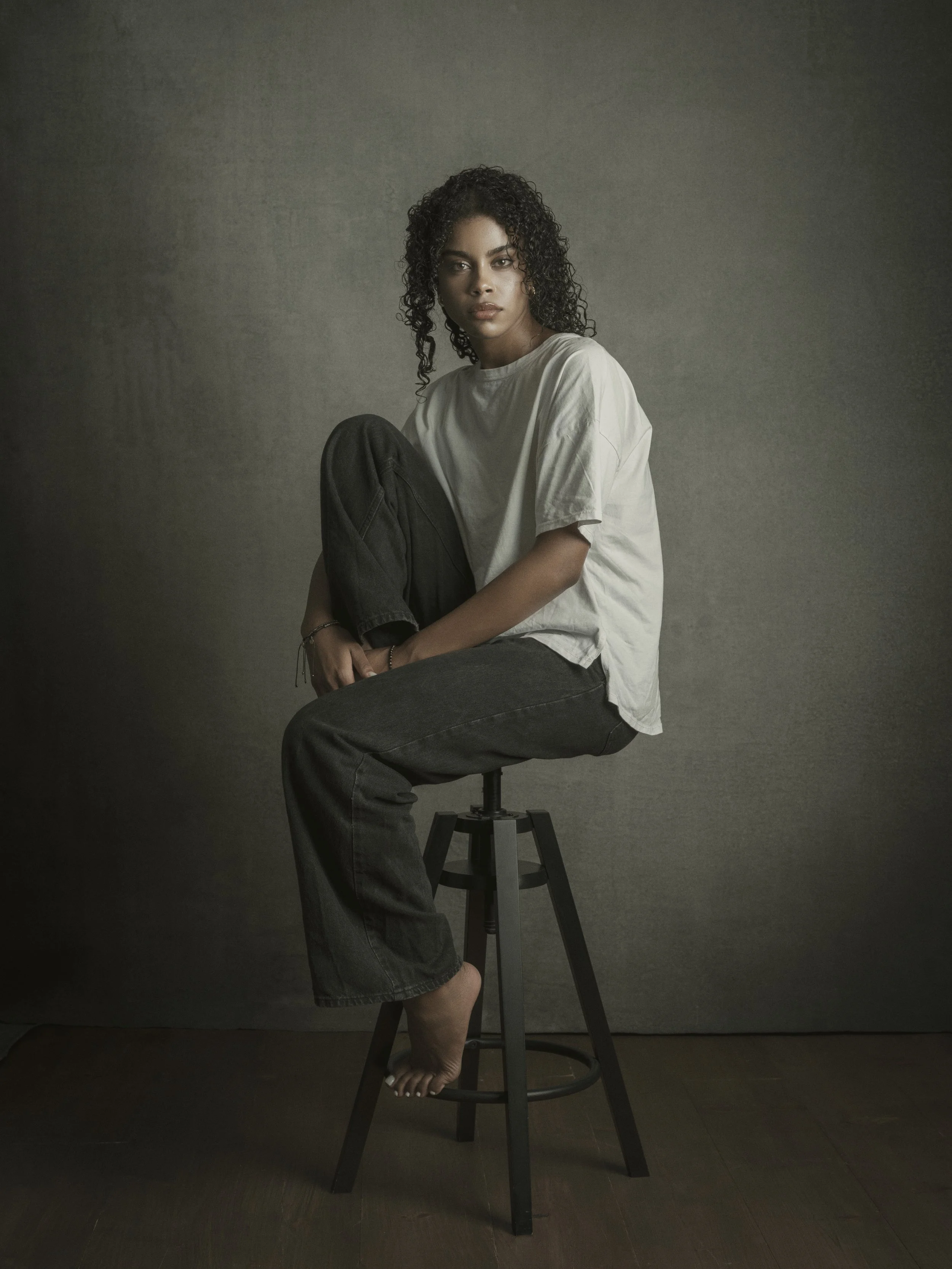 A young woman with curly dark hair and medium skin tone seated on a black stool against a textured gray background. She is wearing a loose white t-shirt and black jeans, with one knee raised close to her chest and her foot resting on the stool's rung