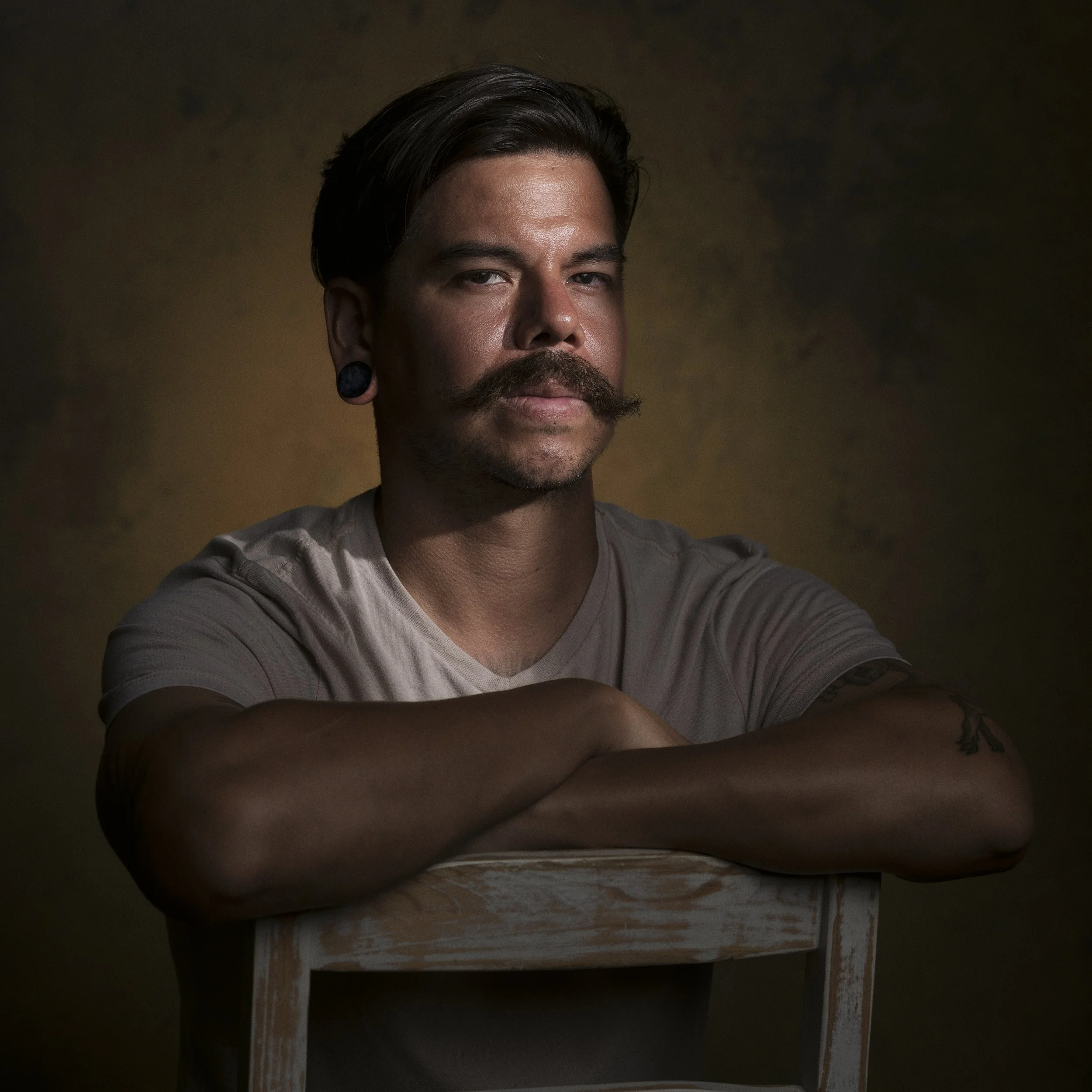 A man with dark hair and a mustache is sitting at a wooden table, looking at the camera, in a dimly lit room.
