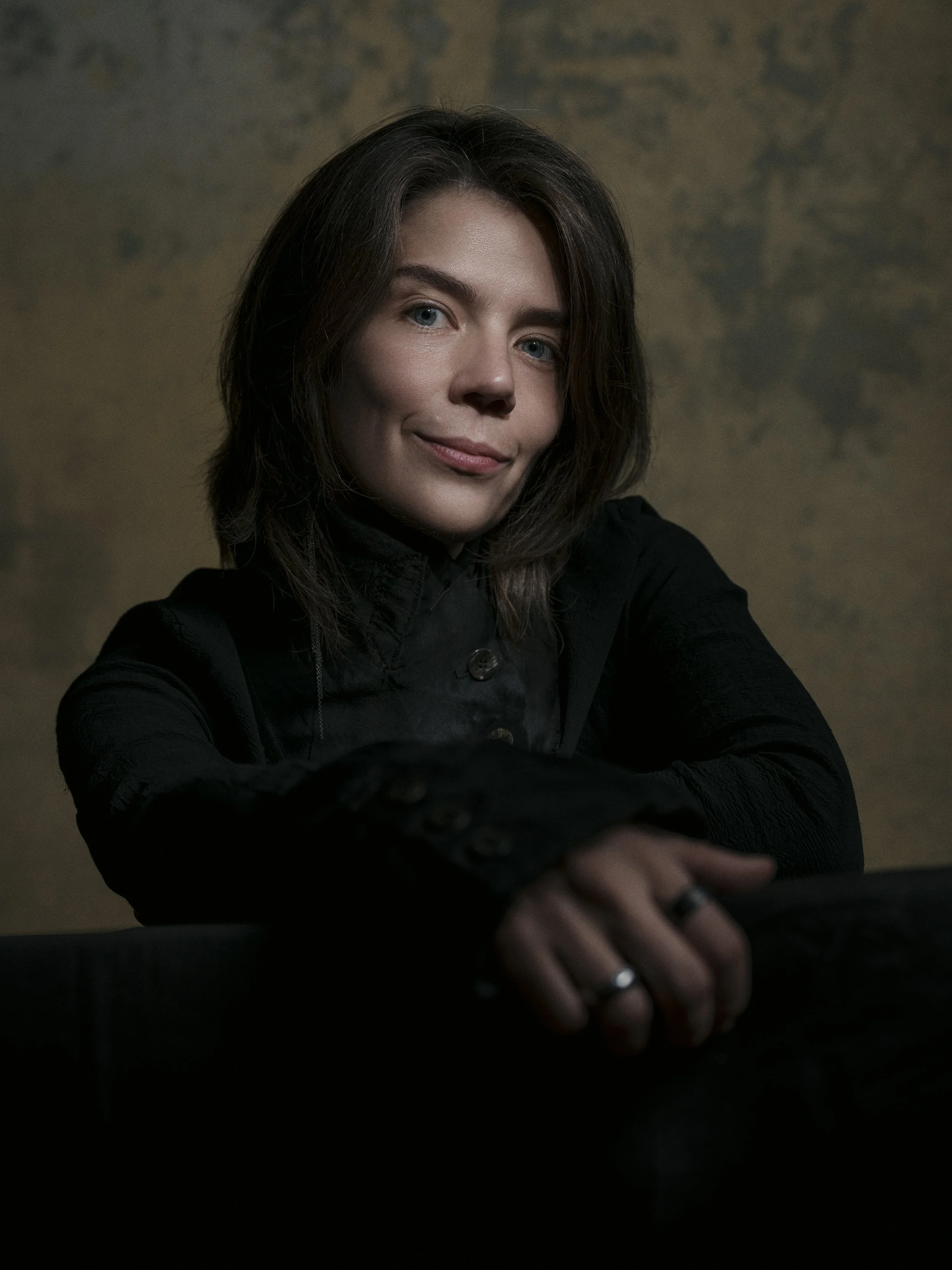 A woman with shoulder-length brown hair and blue eyes in a black jacket, smiling softly, with her hands resting on a dark surface.