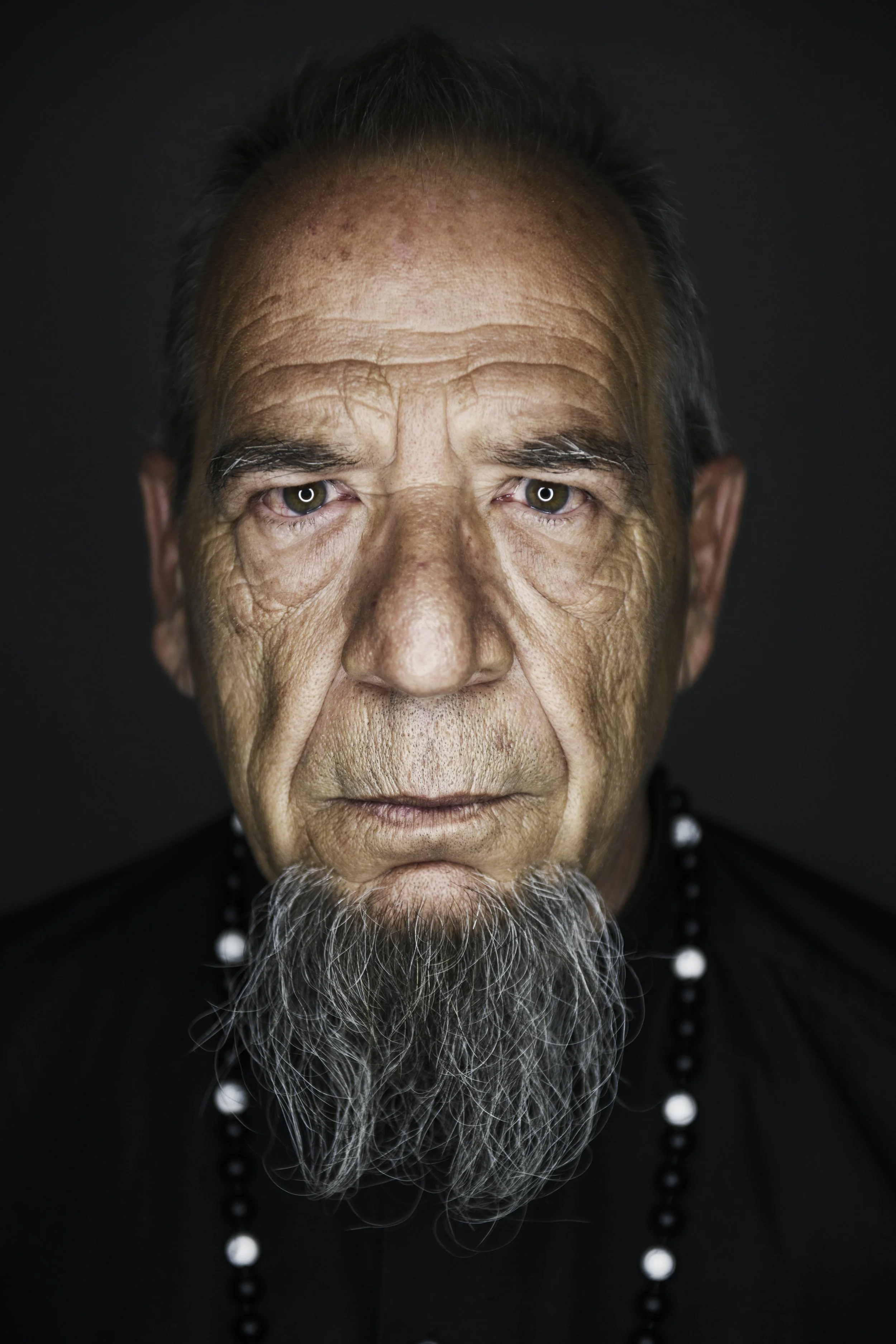 Close-up portrait of an elderly man with a long gray beard, wearing black clothing and a beaded necklace, with a dark background.