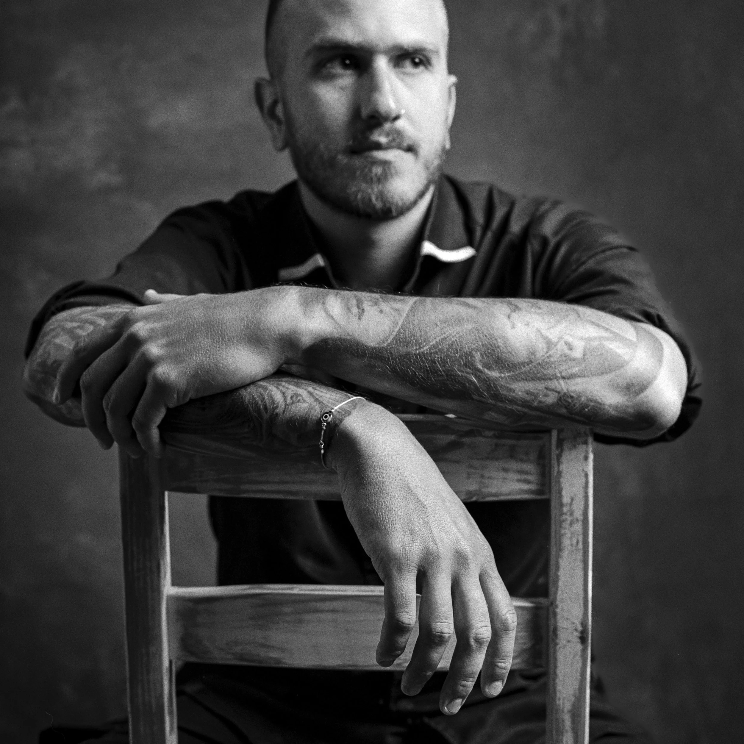 A black and white photo of a man with a beard and short hair, resting his forearms on a wooden surface, with a serious expression, against a cloudy sky background.