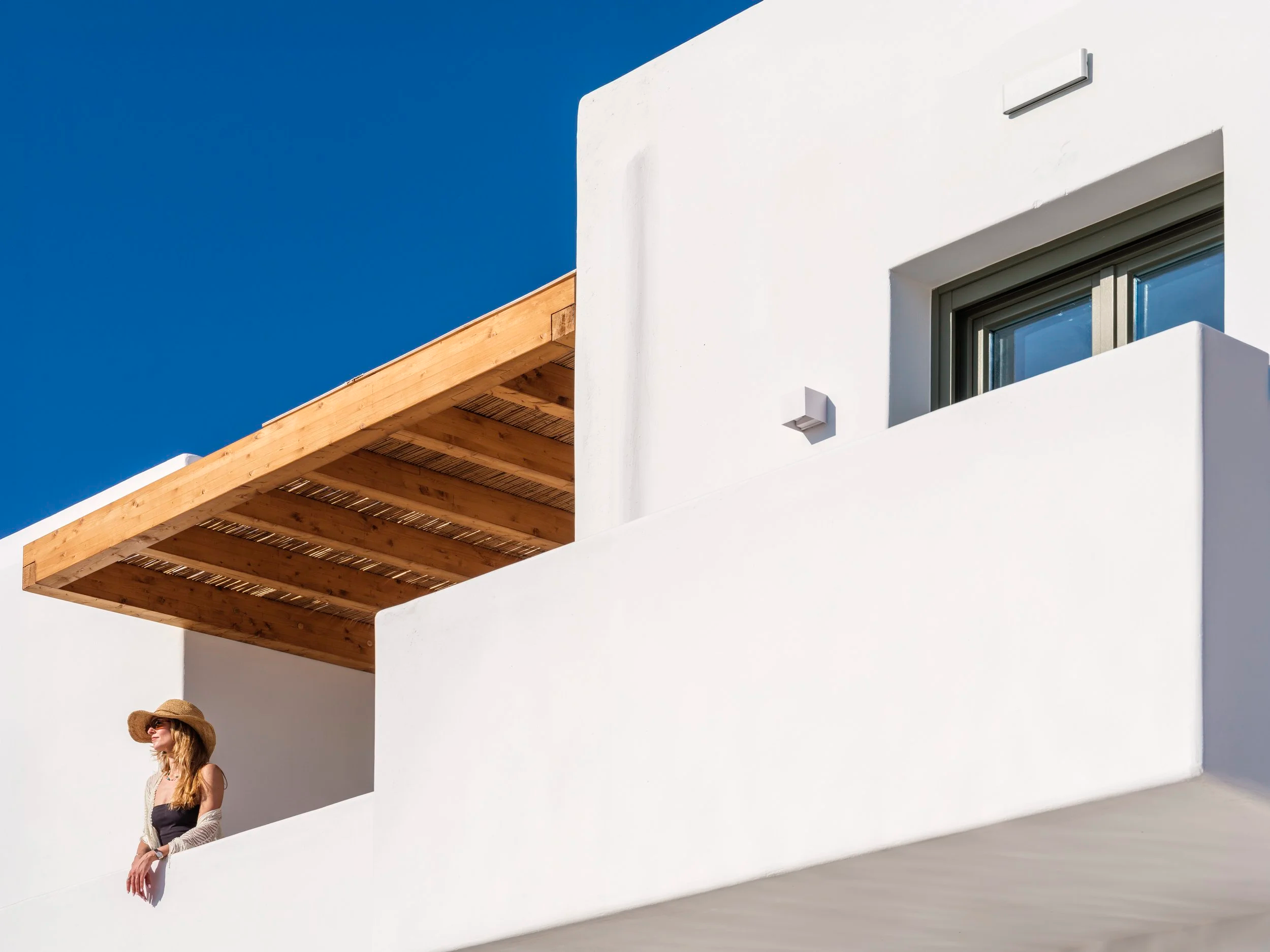 A woman wearing a straw hat and sunglasses stands against the white wall of a modern building with a wooden balcony, under a clear blue sky.